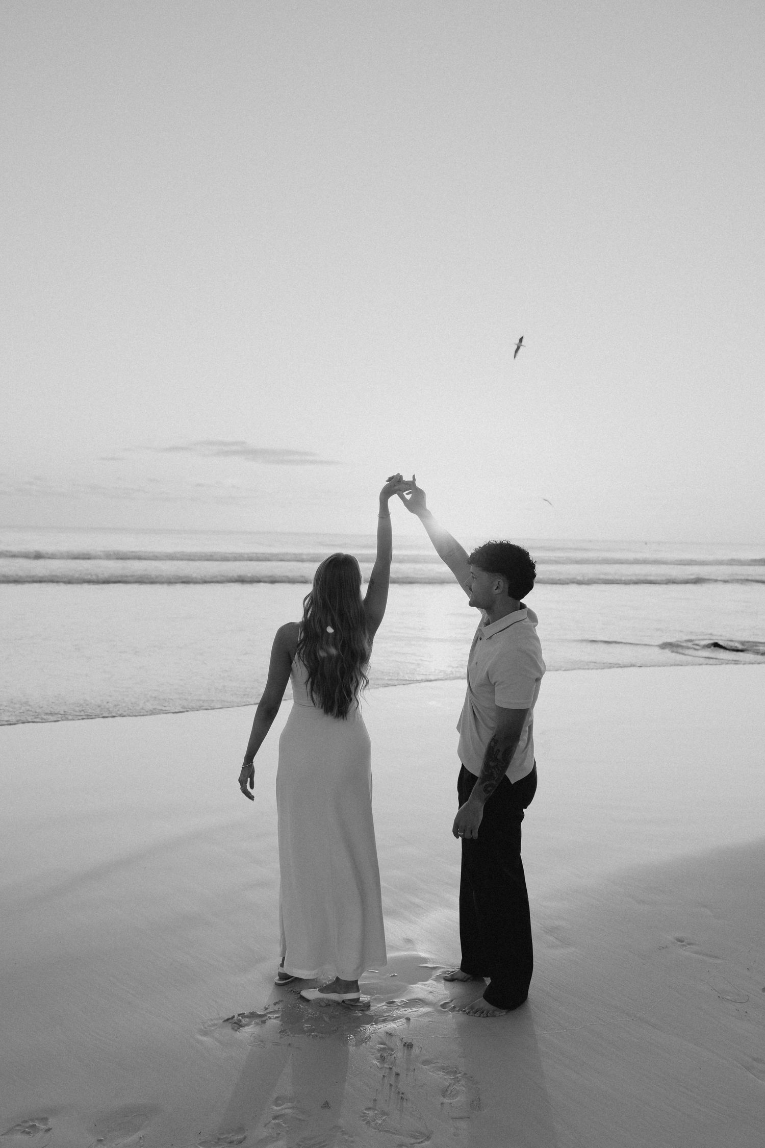 Black and white photo of couple dancing on beach at sunset, bride in white gown.