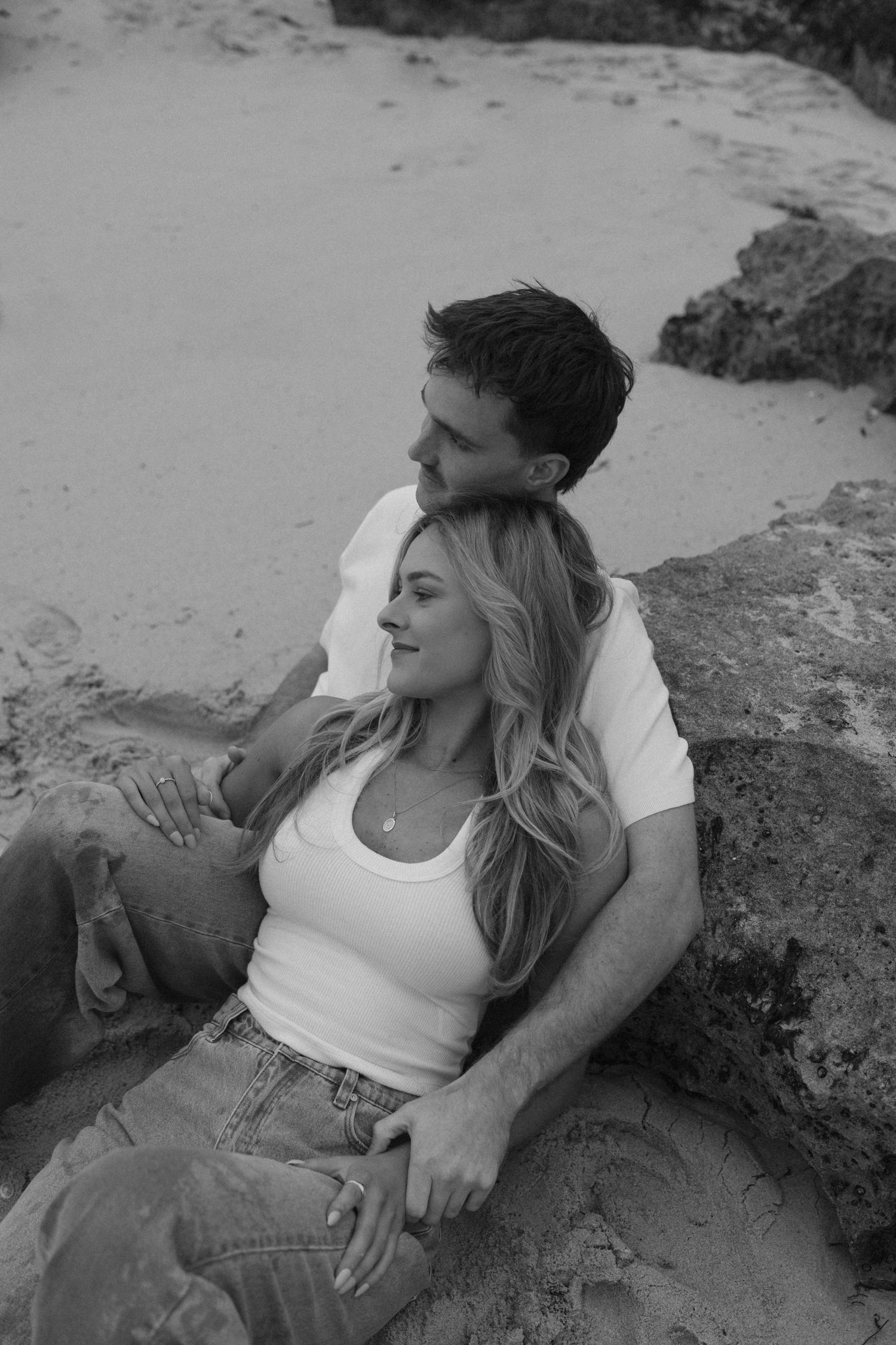 Black and white photo of a couple sitting together on beach rocks, woman leaning back on man.
