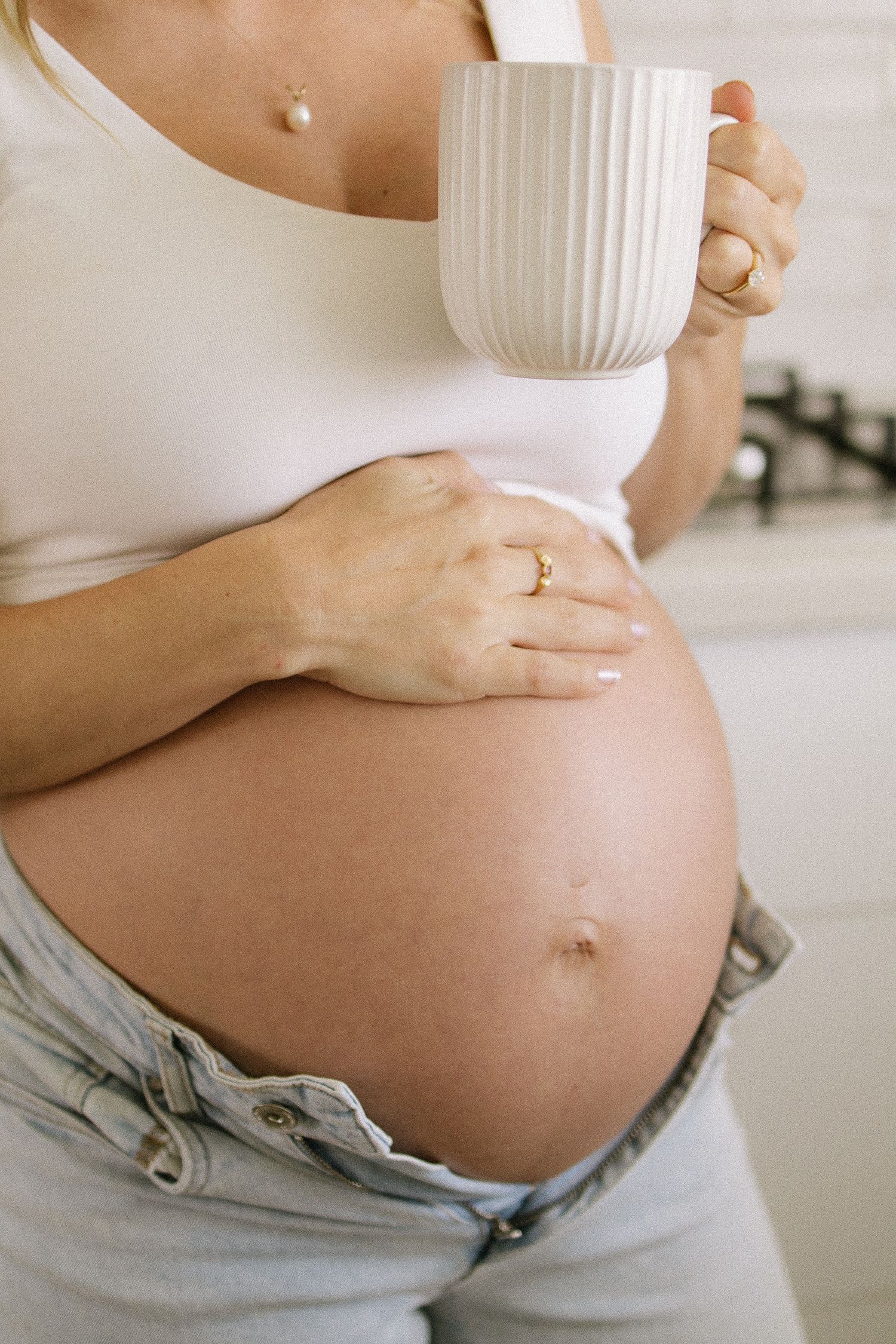 Close-up of pregnant woman holding white ribbed mug while cradling bare baby bump in kitchen.