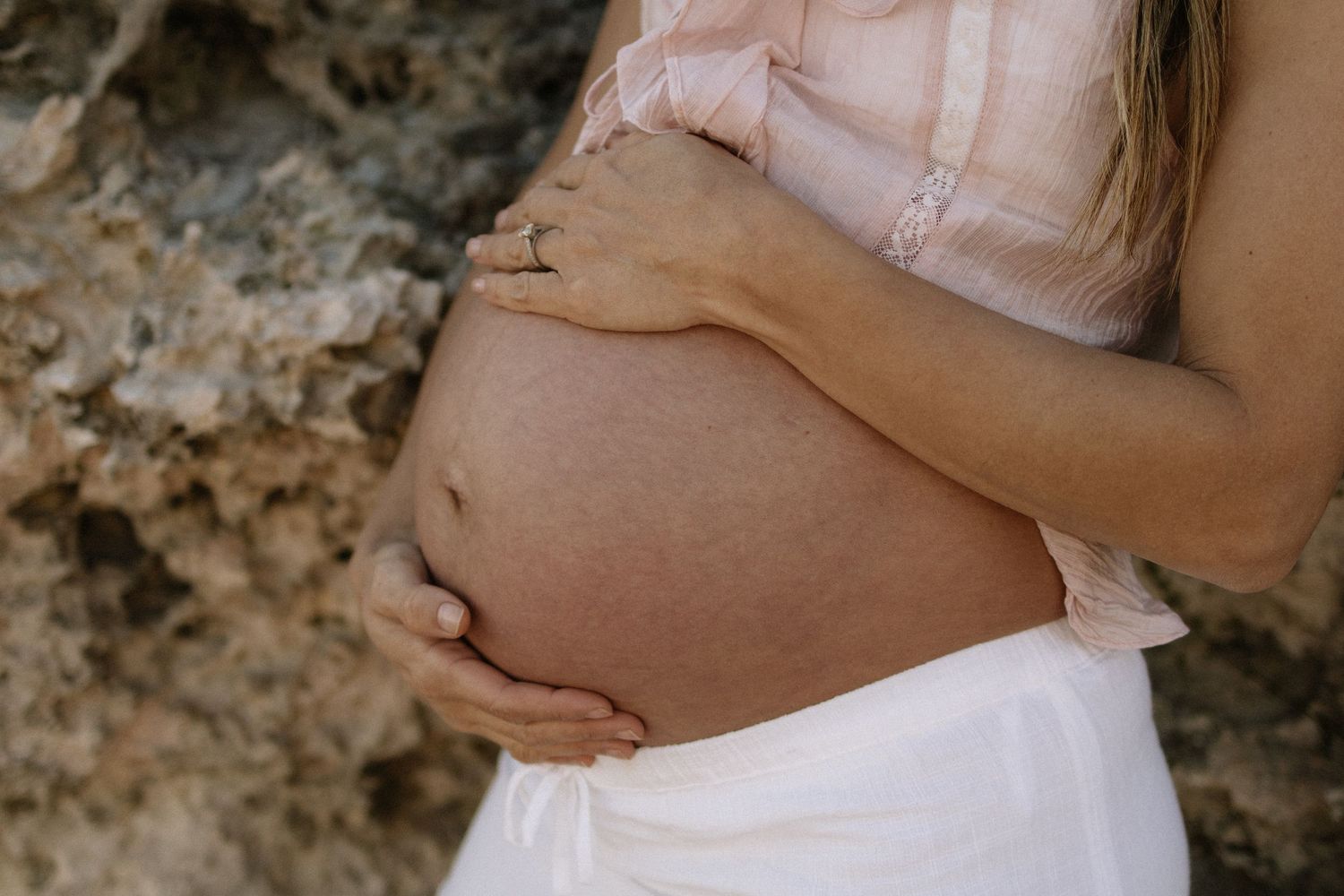Close-up of a pregnant woman cradling her bare belly with both hands near rocky terrain outdoors.