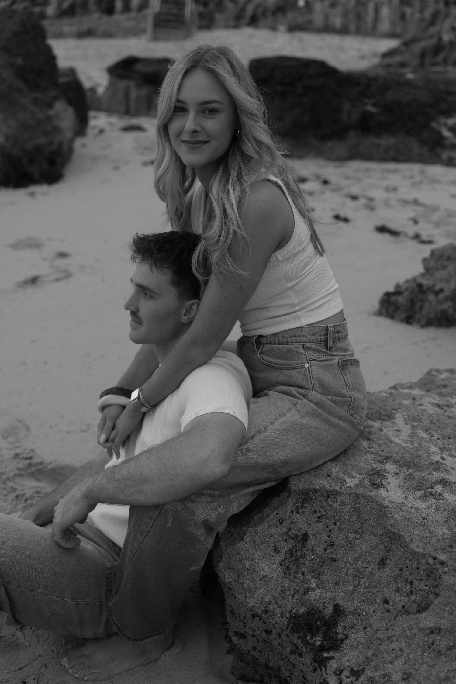 Black and white photo of a couple posing on a rock at the beach, woman sitting behind man smiling.