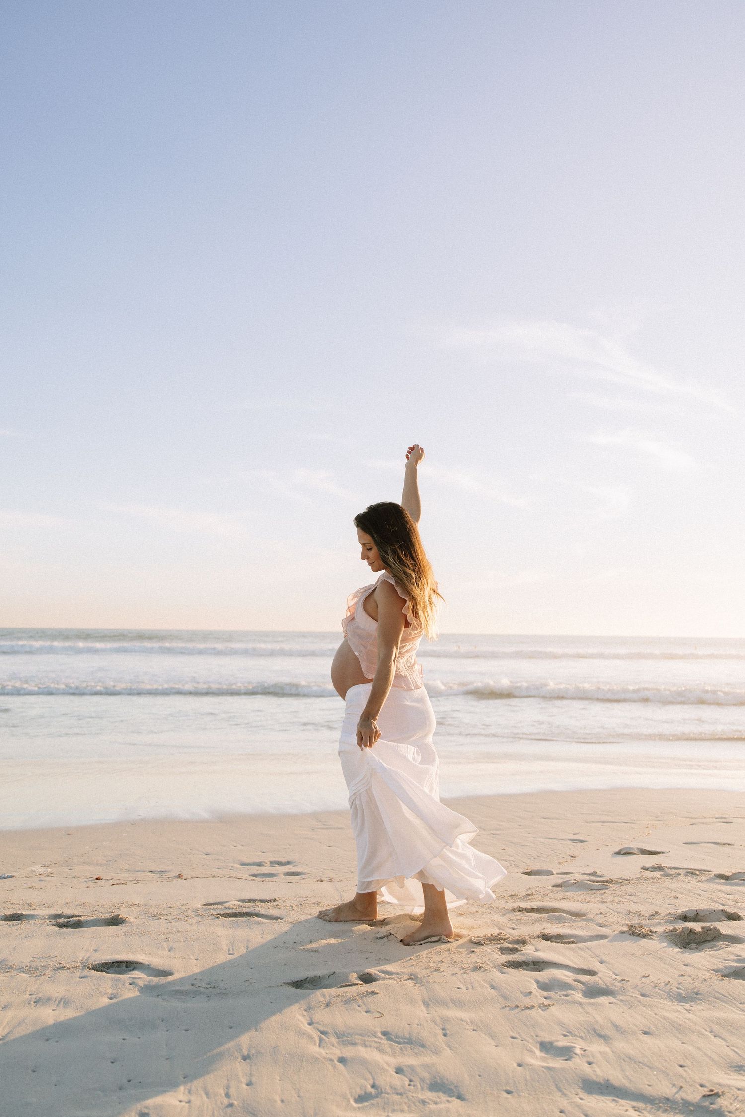 Pregnant woman in flowing white dress raising arm joyfully on beach at sunset.