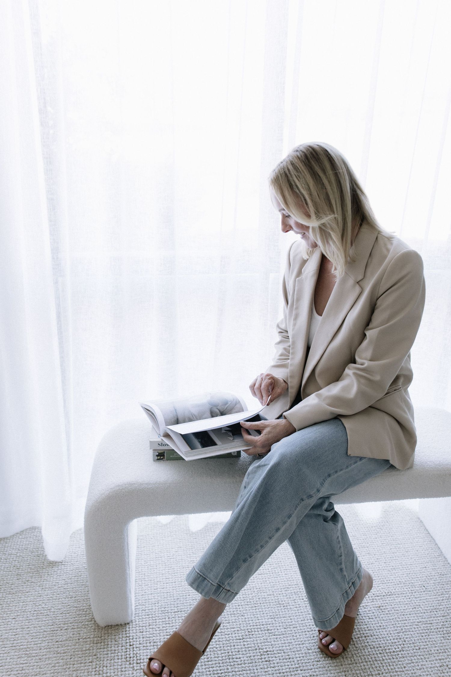 Blonde woman in beige blazer reading a magazine while seated on white bench in bright airy room.