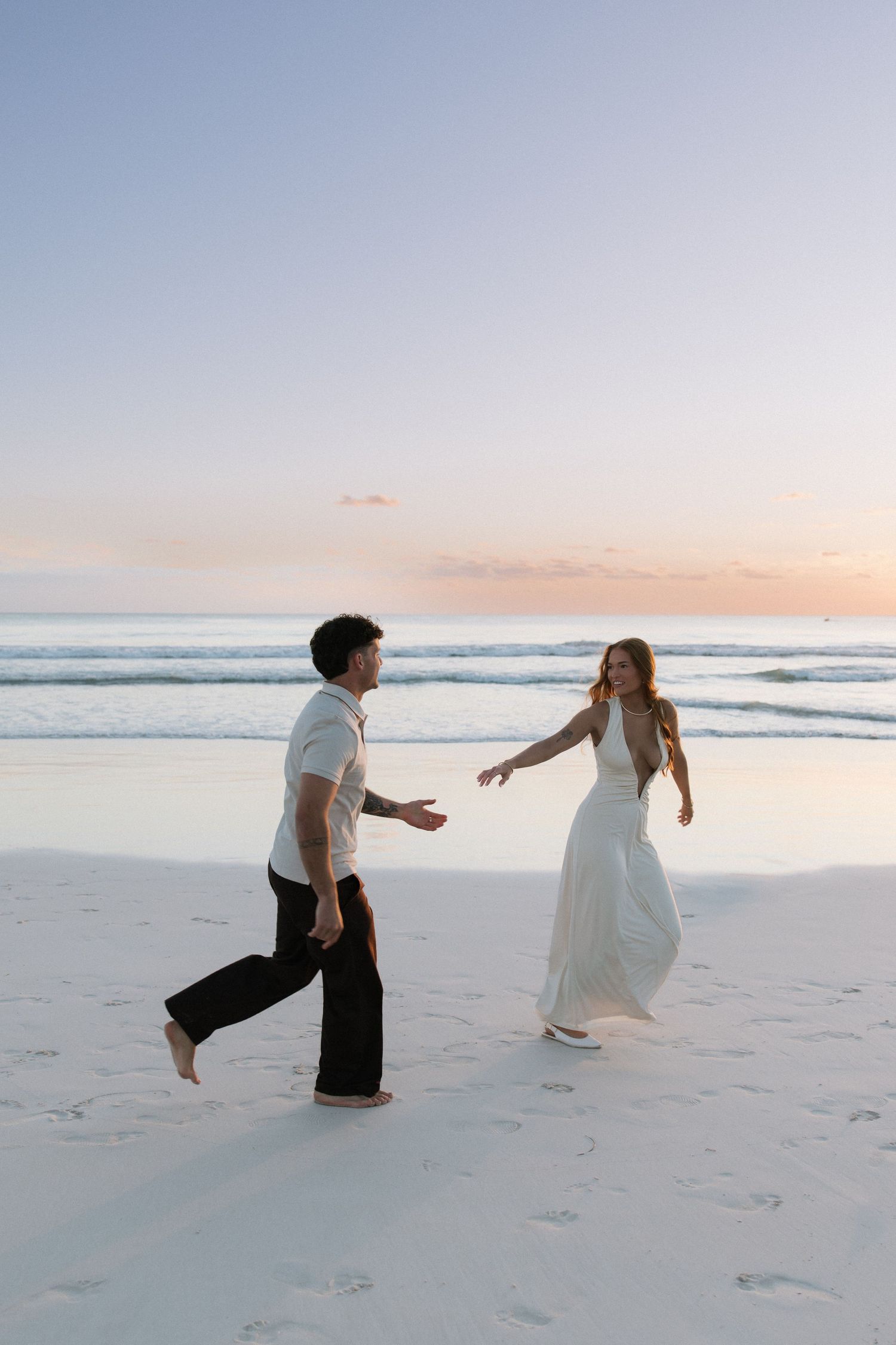 Couple playfully reaching for each other on a wide sandy beach at pastel sunset.