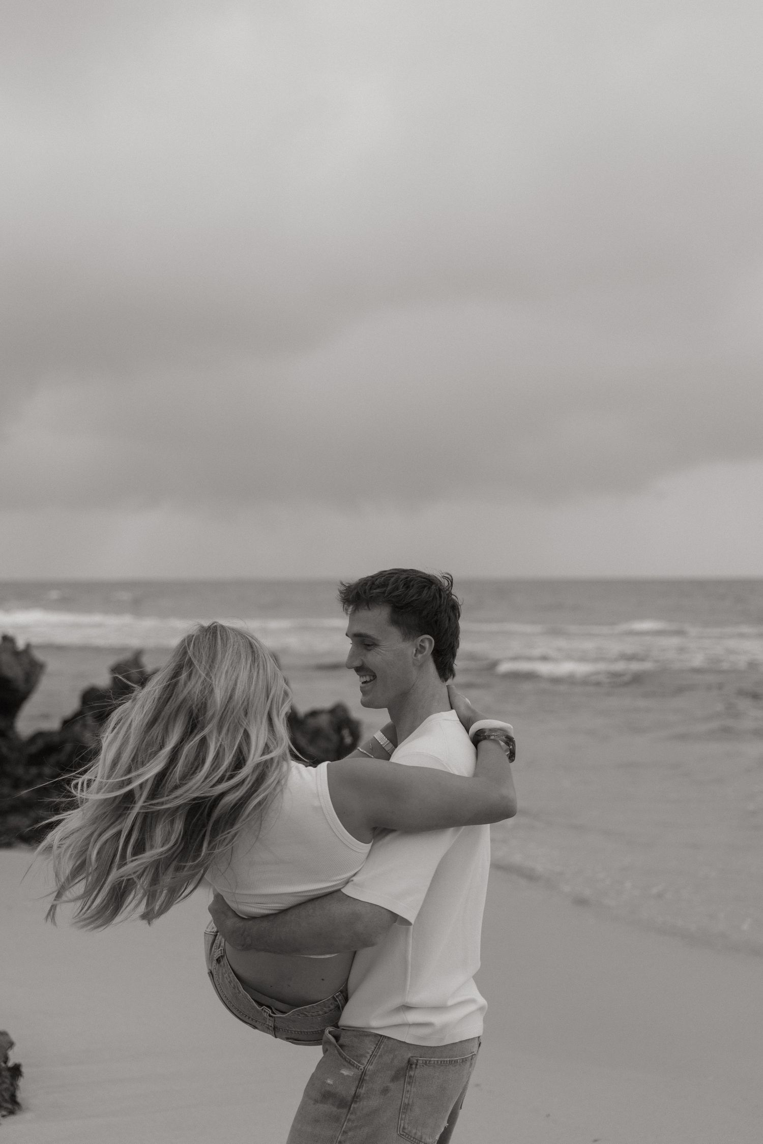 Black and white photo of a couple dancing on a beach with ocean waves and cloudy sky behind them.