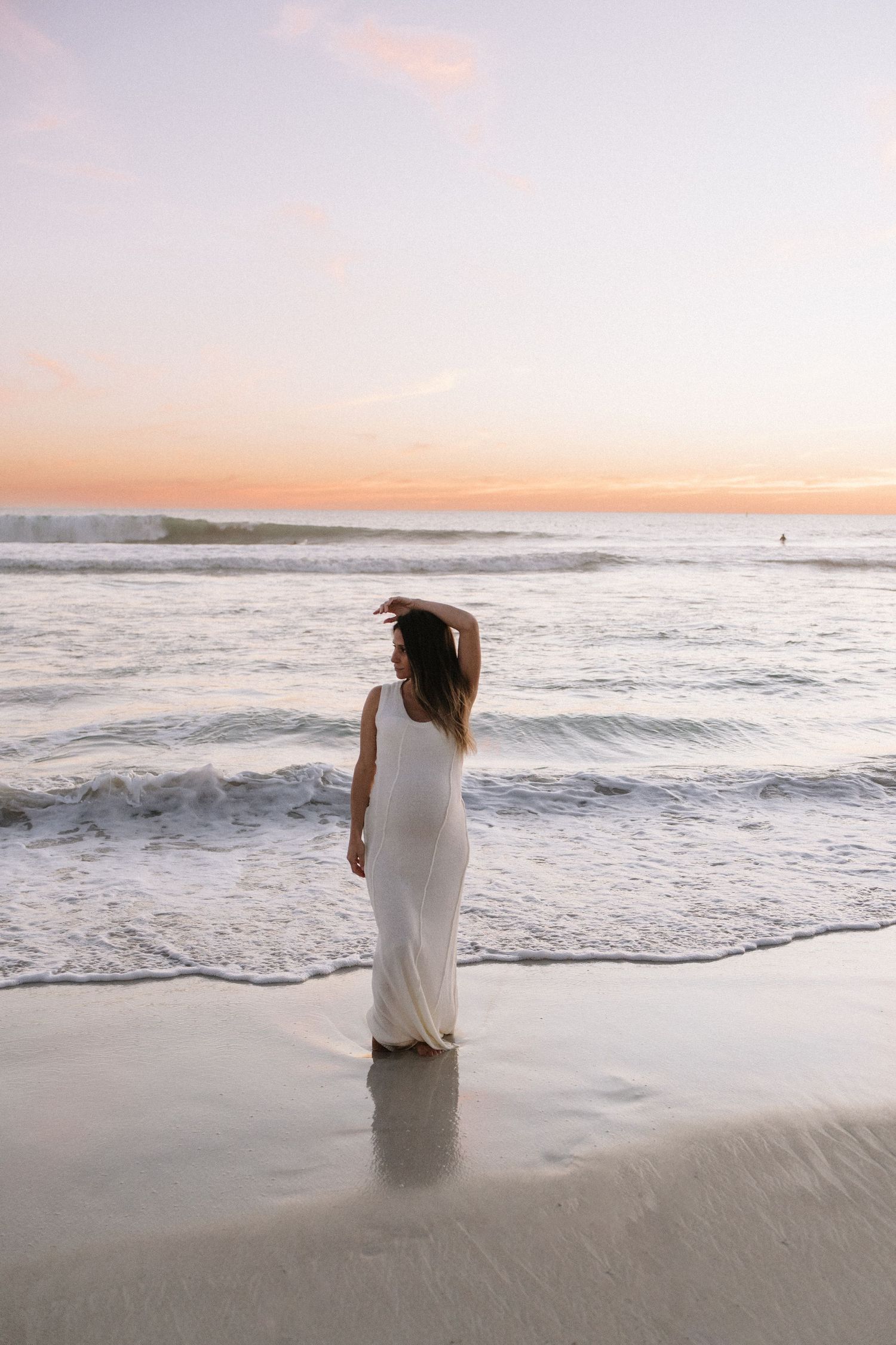 Pregnant woman in white maxi dress standing at ocean's edge at sunset.