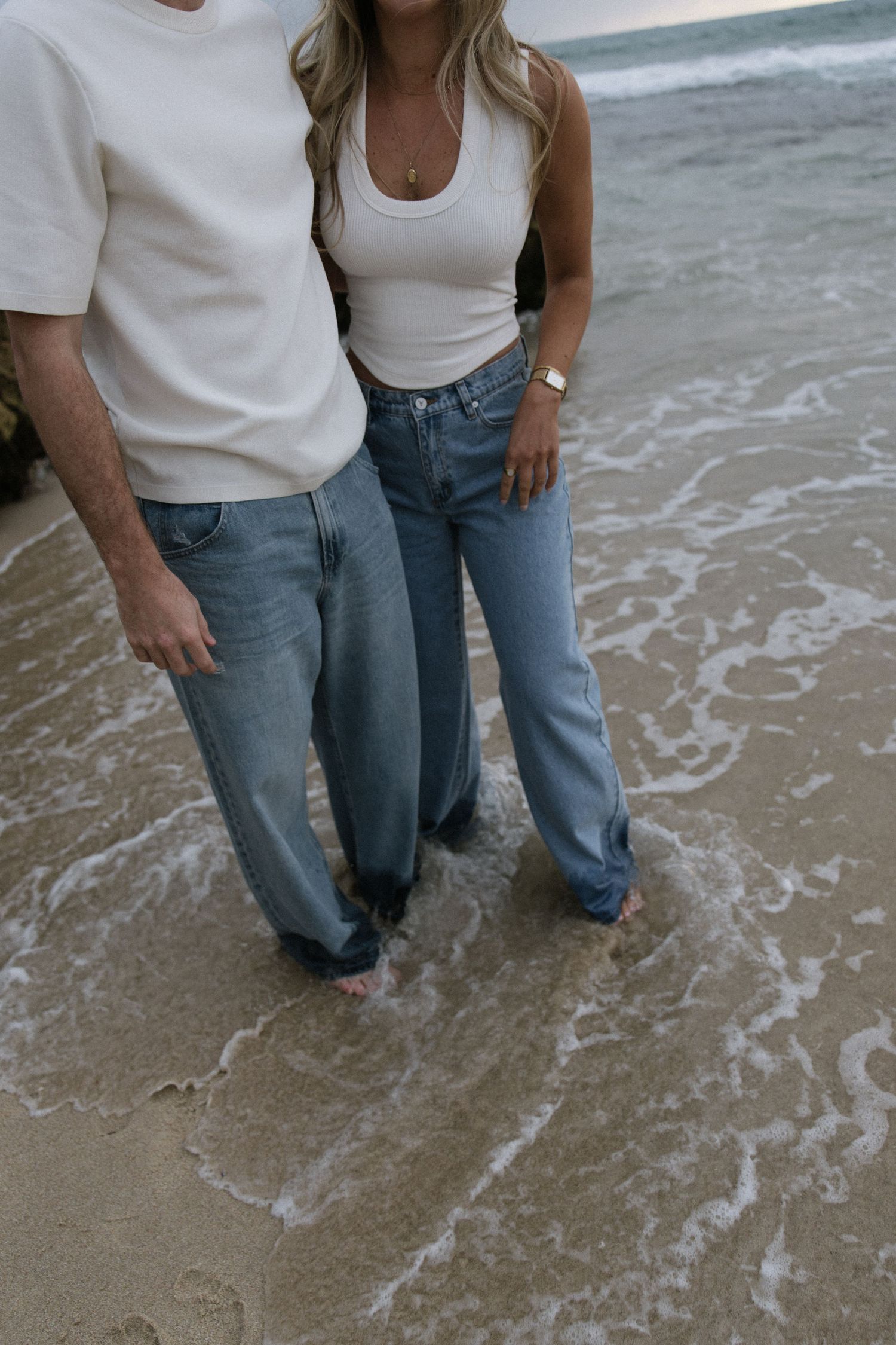 Couple standing barefoot in ocean waves on a sandy beach wearing white tops and blue jeans.