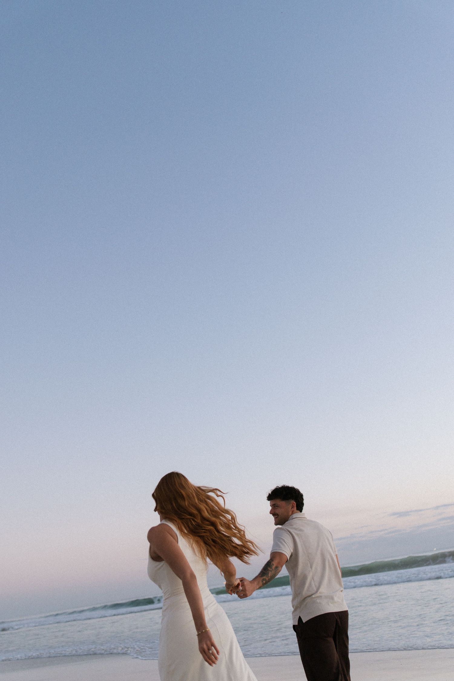Woman in white dress leads man by hand along beach during golden hour sunset.