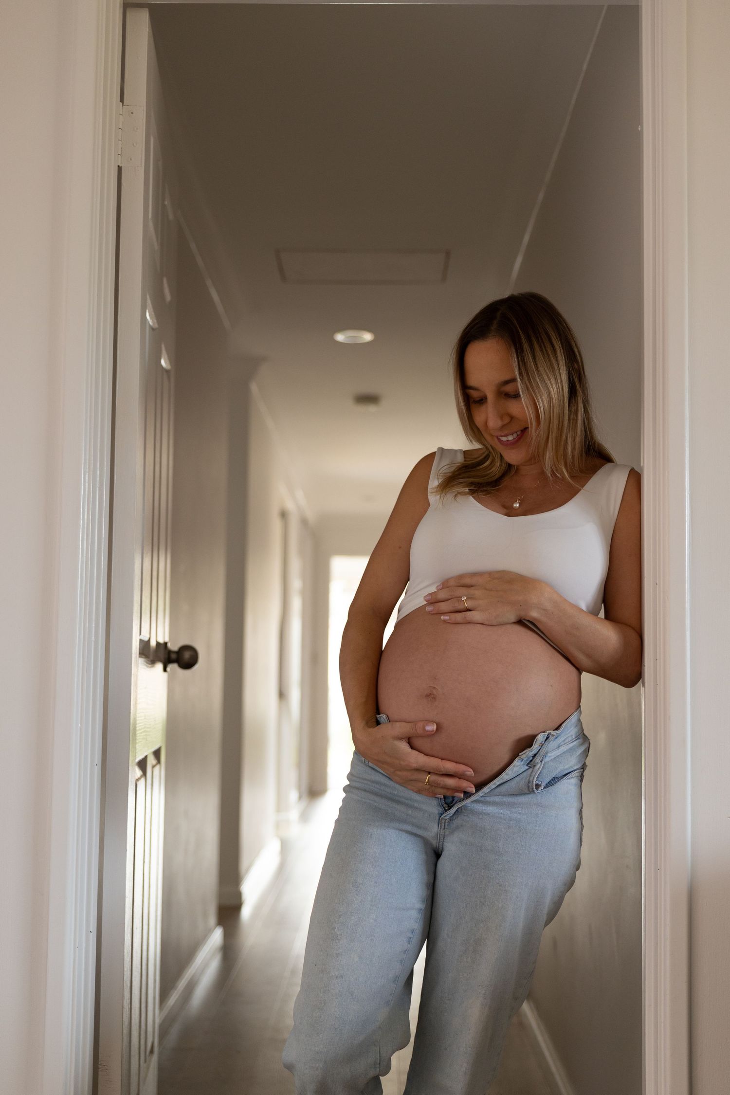Pregnant woman in white crop top and jeans smiling at baby bump while leaning in sunlit hallway.