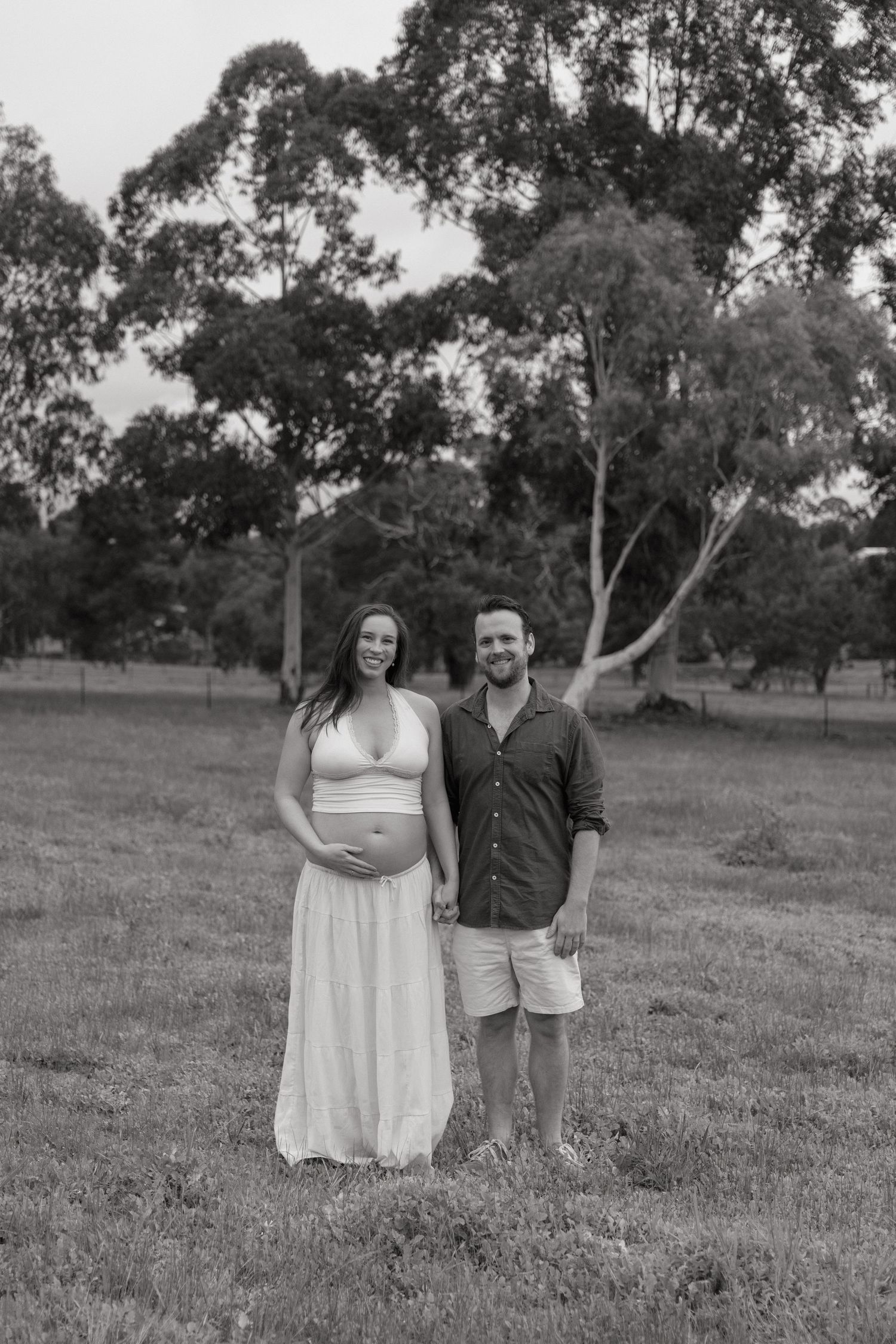Black and white maternity photo of a pregnant woman and partner holding hands in a grassy park.