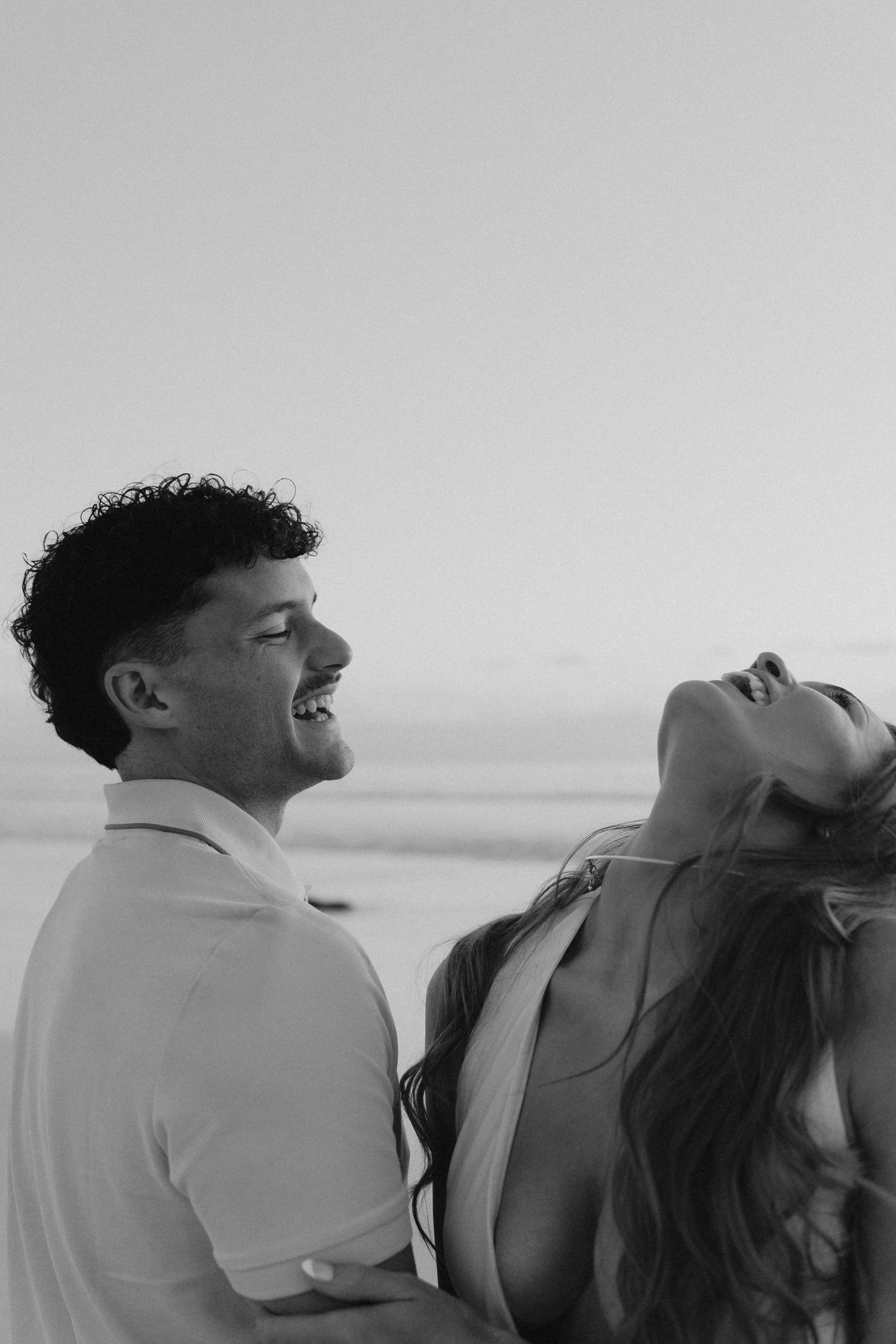 Black and white photo of laughing couple embracing on beach during romantic sunset session.