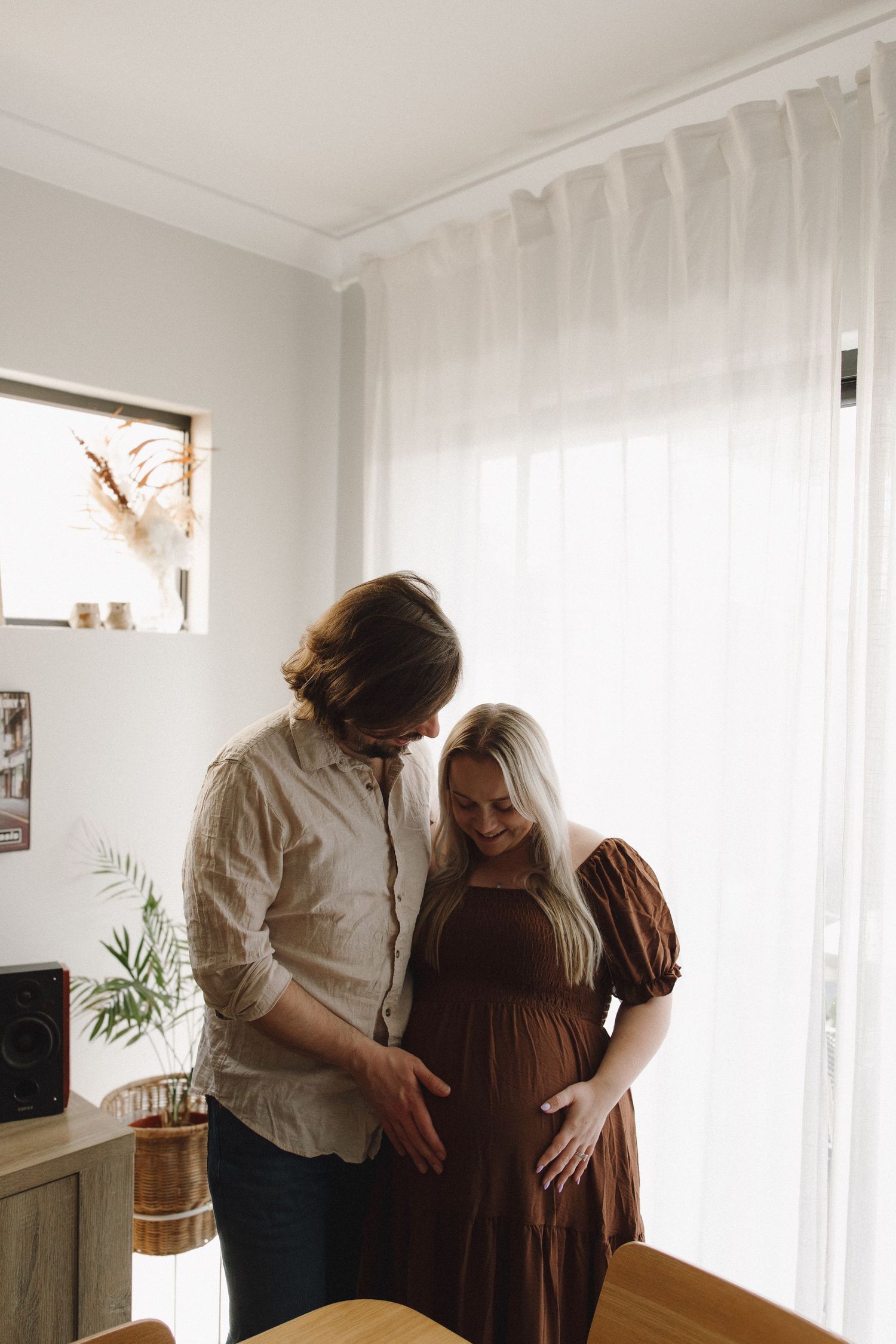 Couple holding pregnant belly together near large sunlit window indoors.