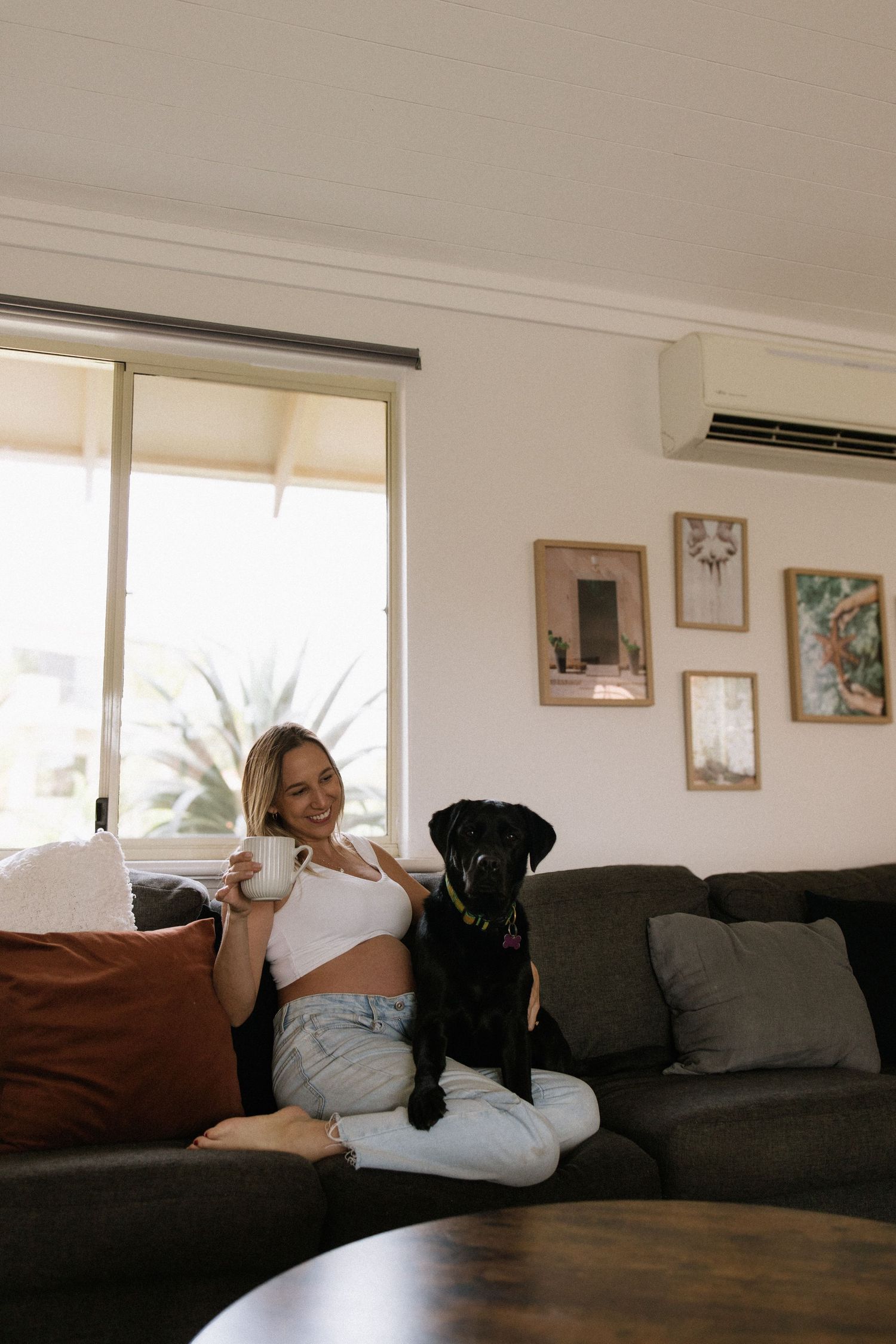 Smiling pregnant woman sitting on a couch with a black Labrador, holding a white mug in a cozy living room.