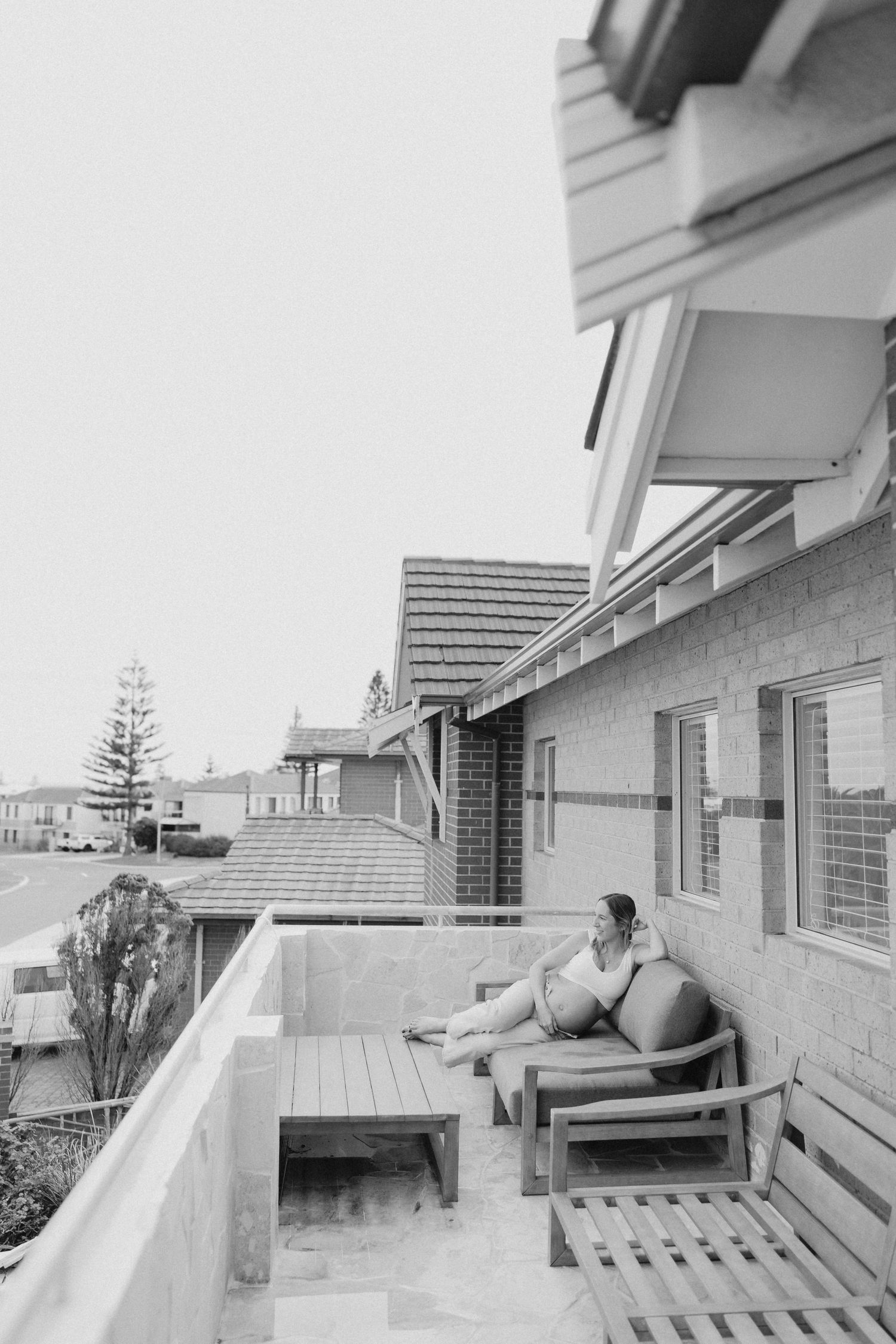 A person relaxes on outdoor furniture on a balcony overlooking a suburban street, captured in black and white.