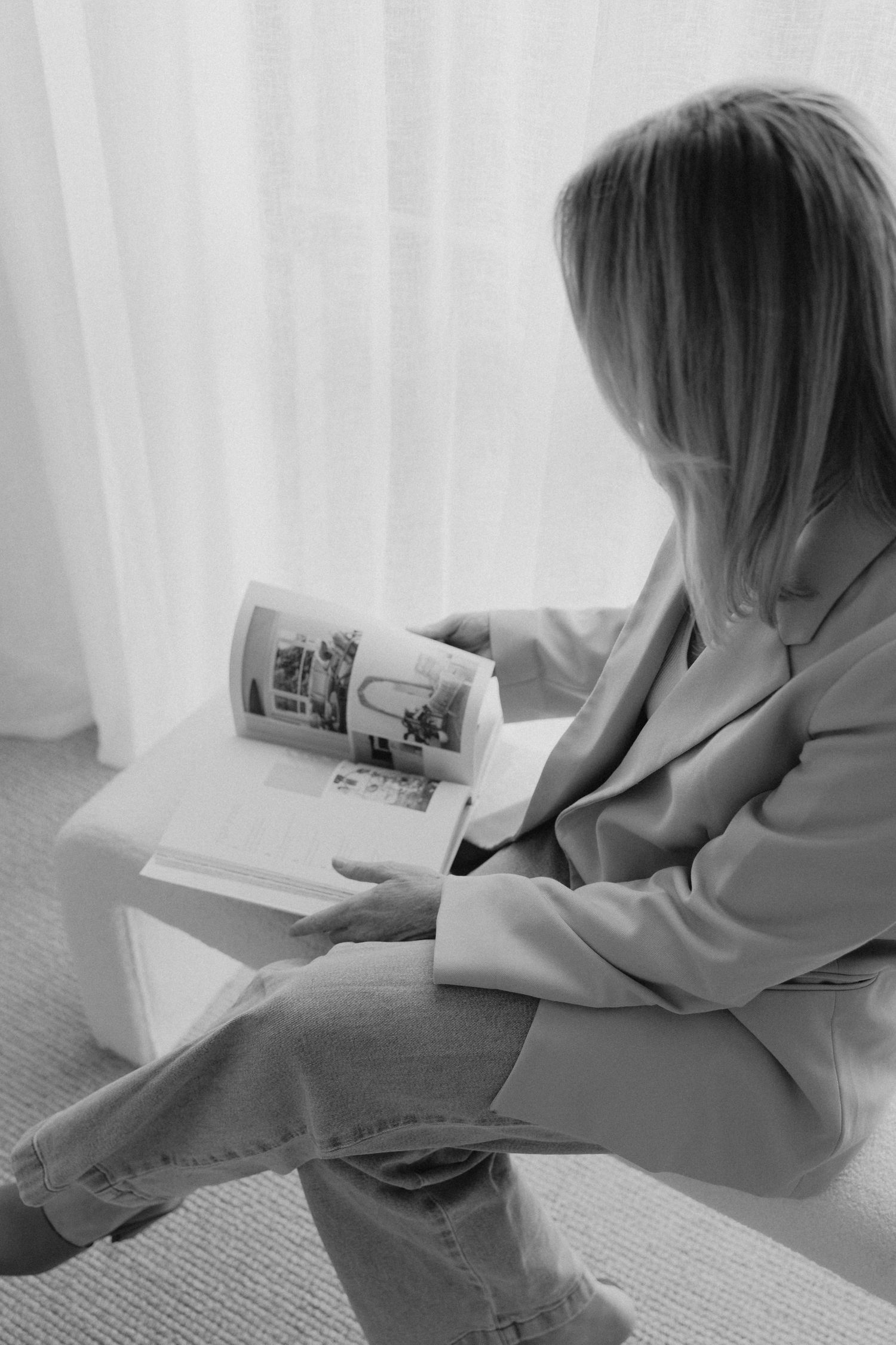 Black and white photo of blonde woman in blazer reading a magazine on a white bench.