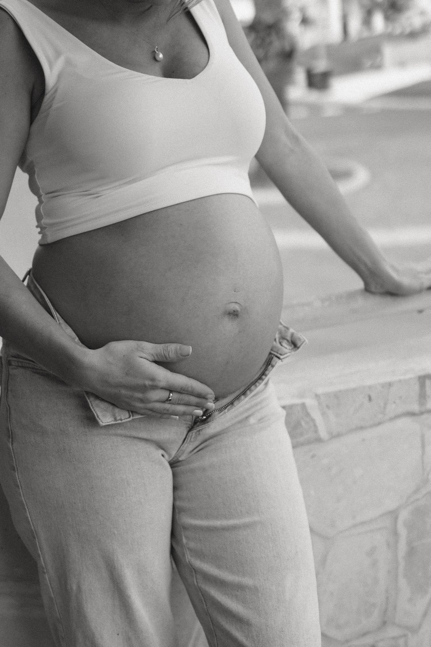 Black and white close-up of pregnant woman in white crop top cradling bare belly outdoors.