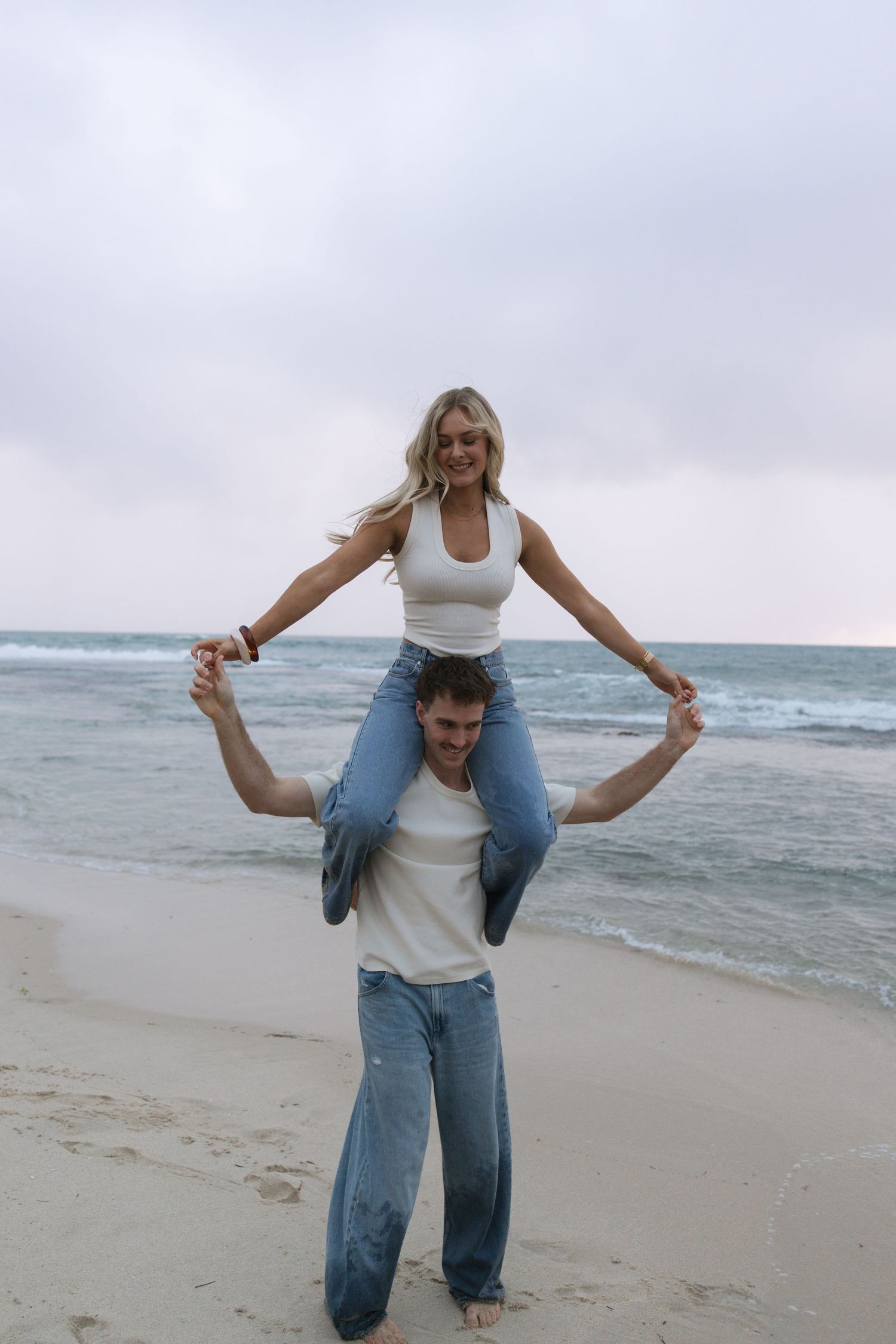 Smiling woman sitting on man's shoulders on a sandy beach with ocean waves in the background.