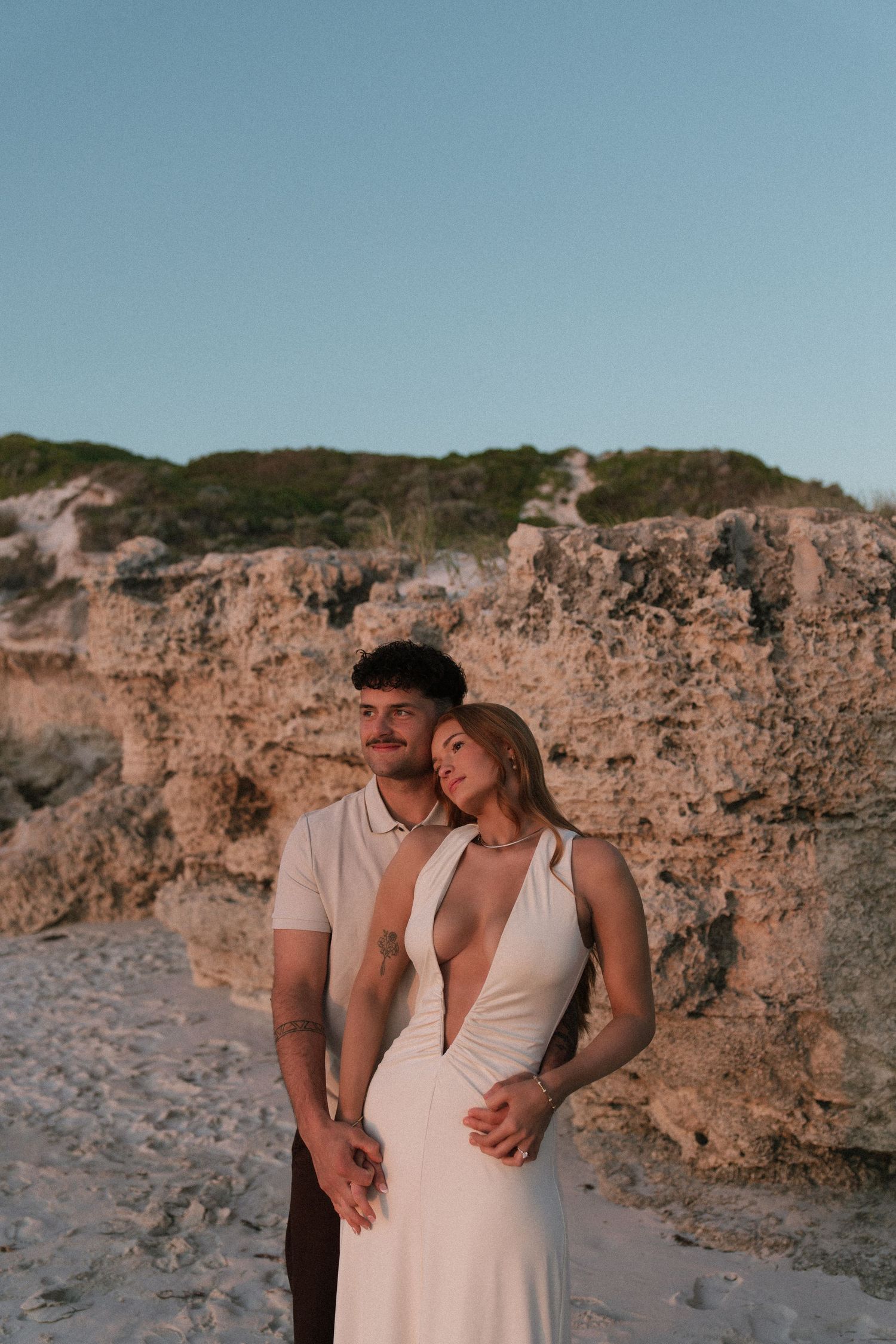 Couple posing together near rocky cliffs on sandy beach at golden hour.
