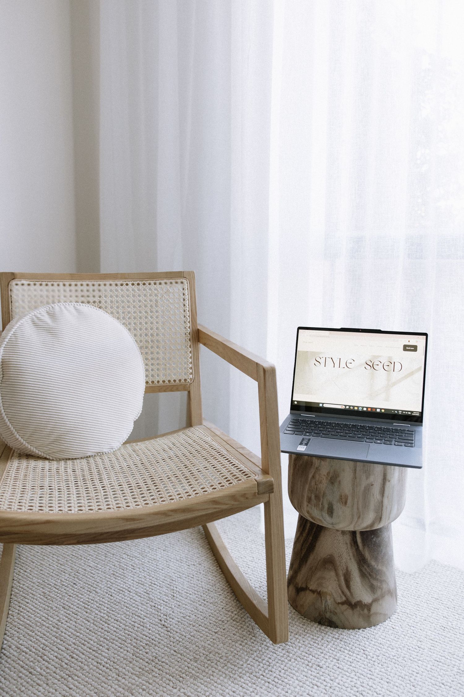 Minimalist rattan rocking chair with white cushion beside laptop on wooden stump side table near sheer curtains.