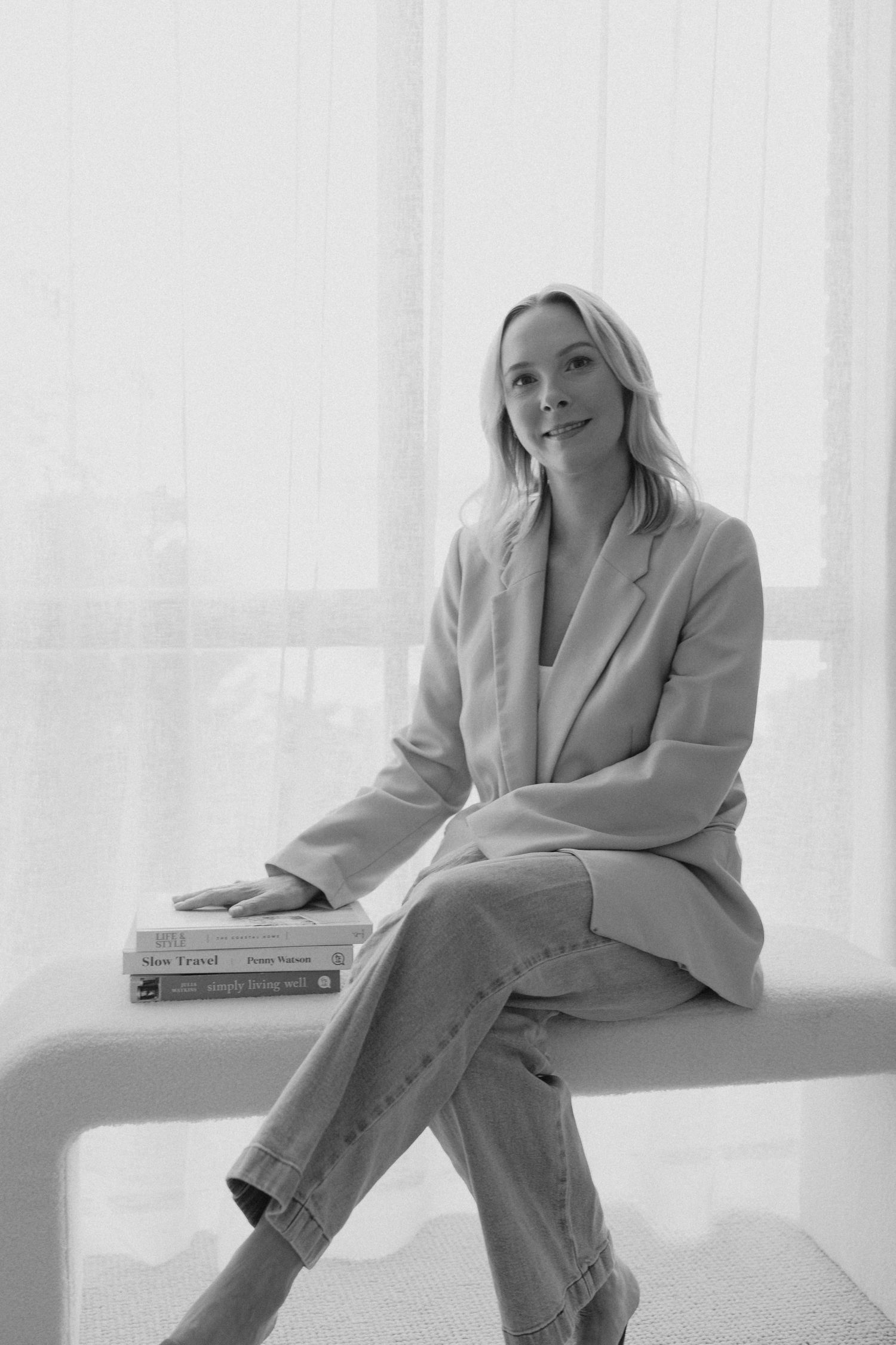 Elegant blonde woman in beige blazer sitting on white bench with stacked books in black and white photo.