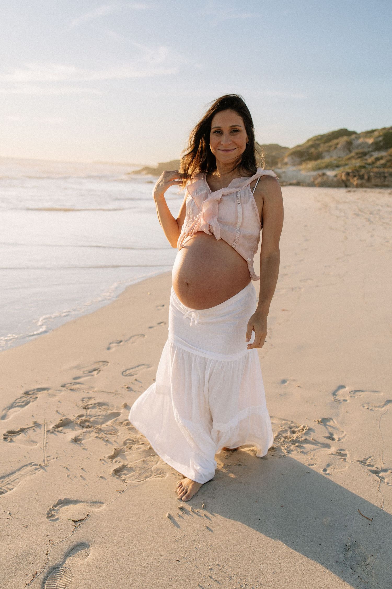 Smiling pregnant woman in pink top and white skirt standing on sunny beach at golden hour.