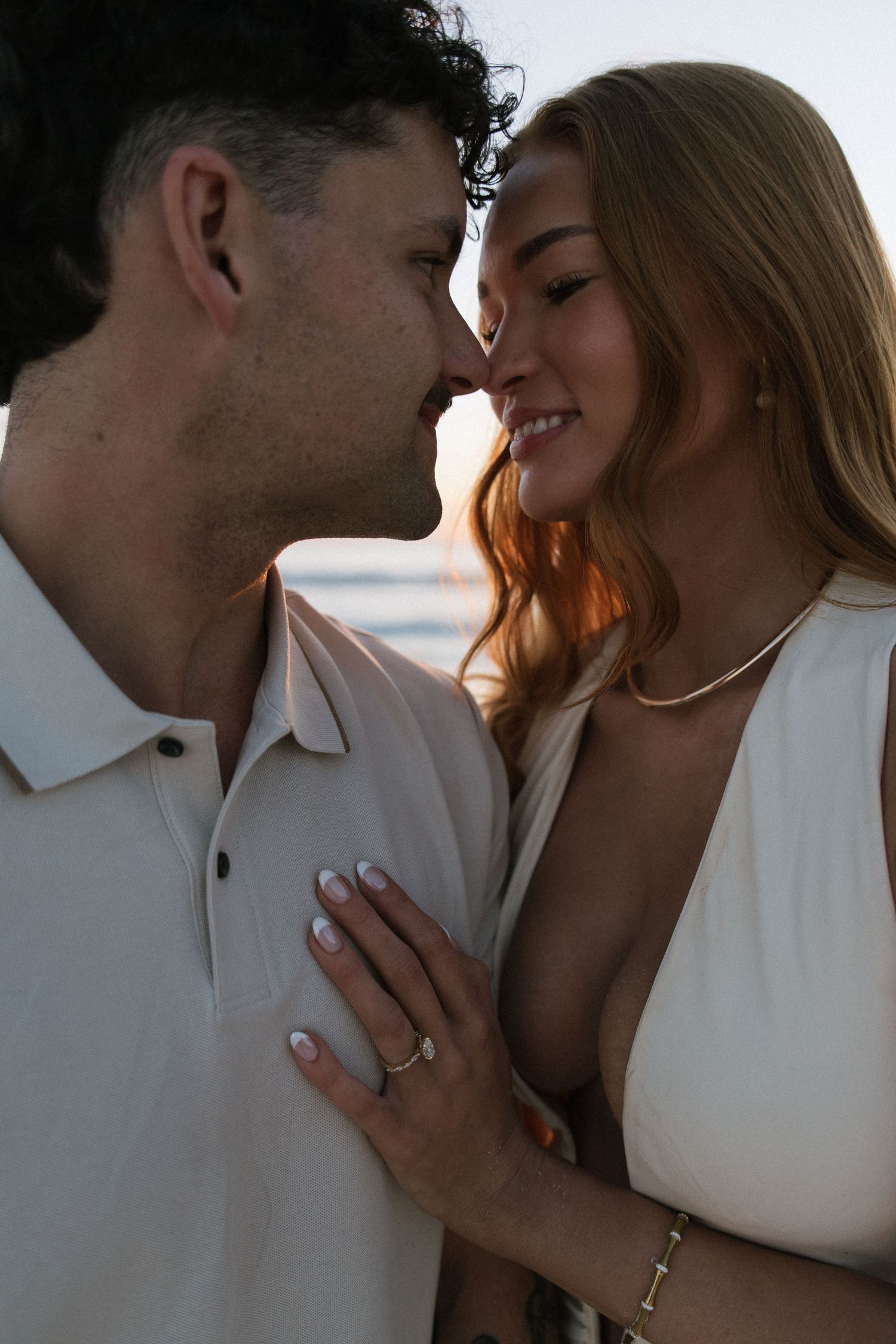 Intimate close-up of couple nearly kissing at sunset beach, woman displaying engagement ring on hand.