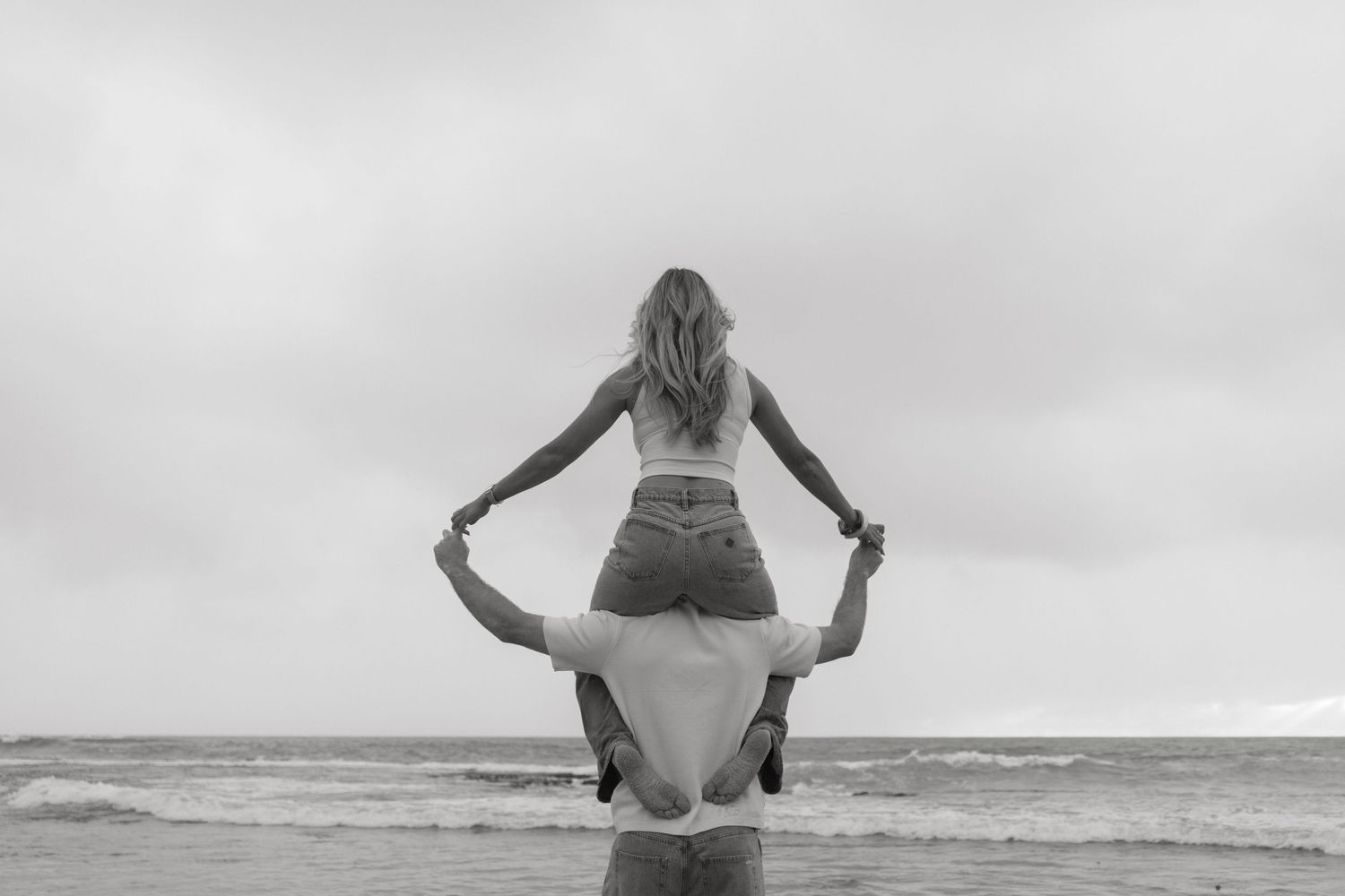 Black and white photo of woman sitting on mans shoulders at ocean beach.