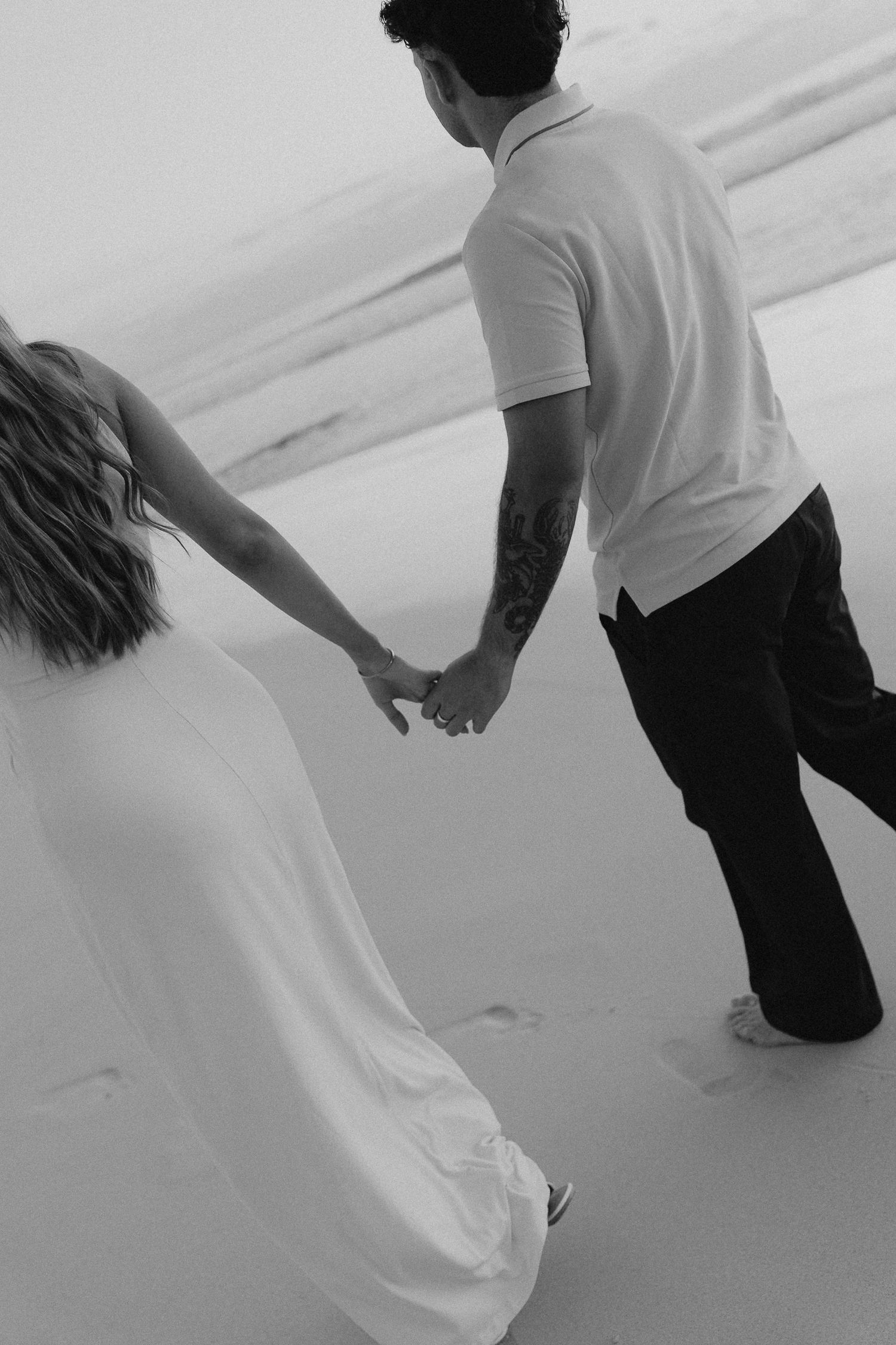 Black and white photo of engaged couple holding hands while walking on sandy beach.