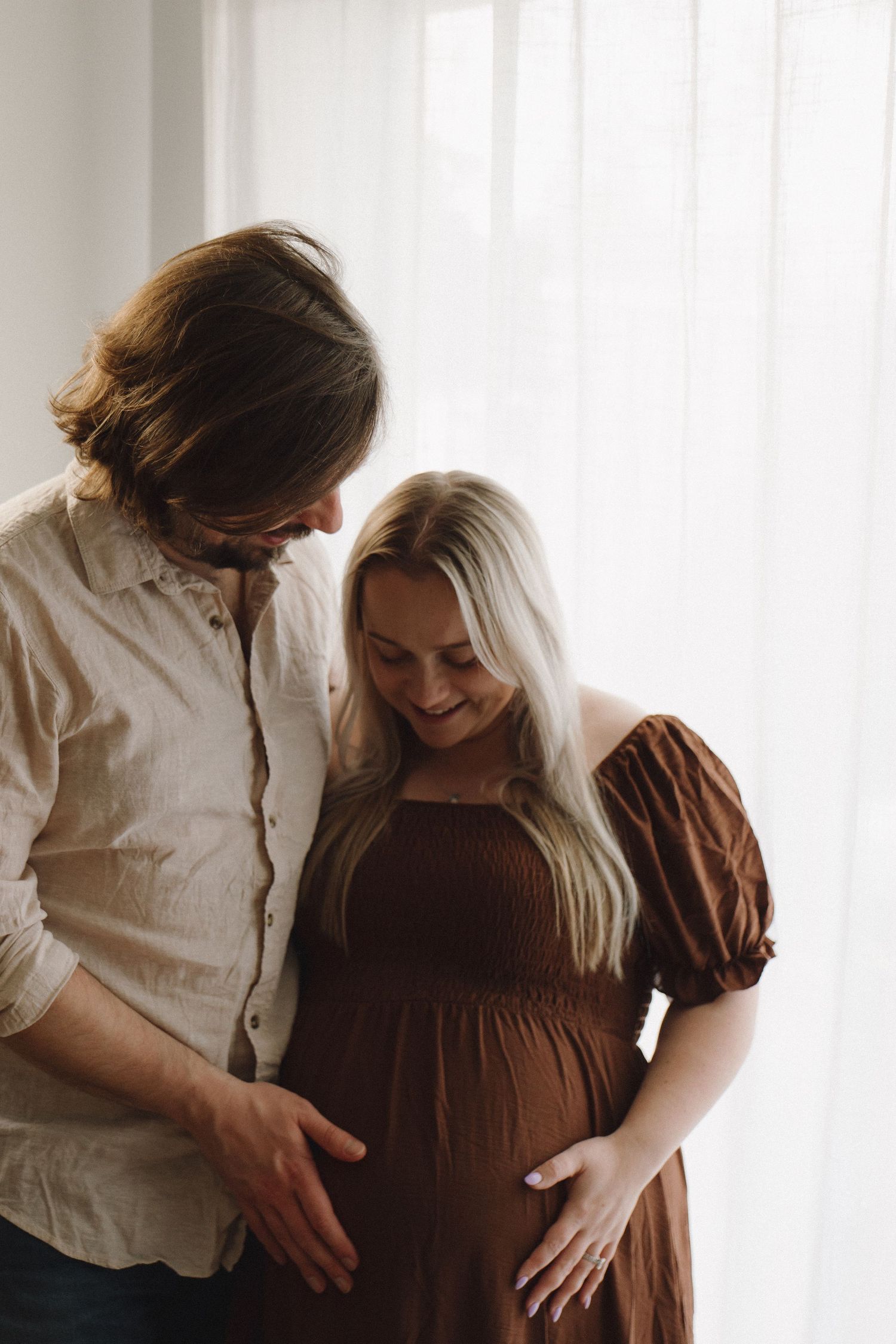Couple smiling and touching pregnant belly indoors by bright window.