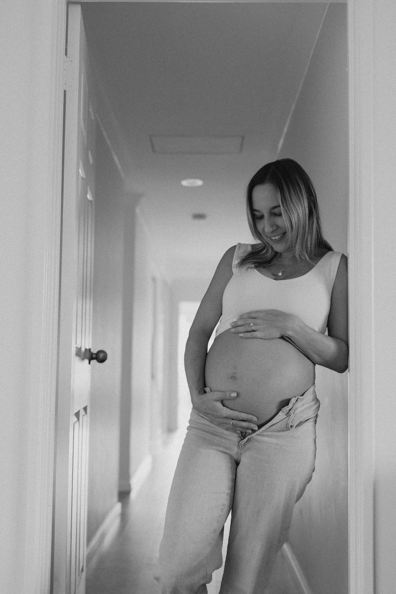 Black and white photo of a smiling pregnant woman in a hallway, gently holding her bare baby bump.