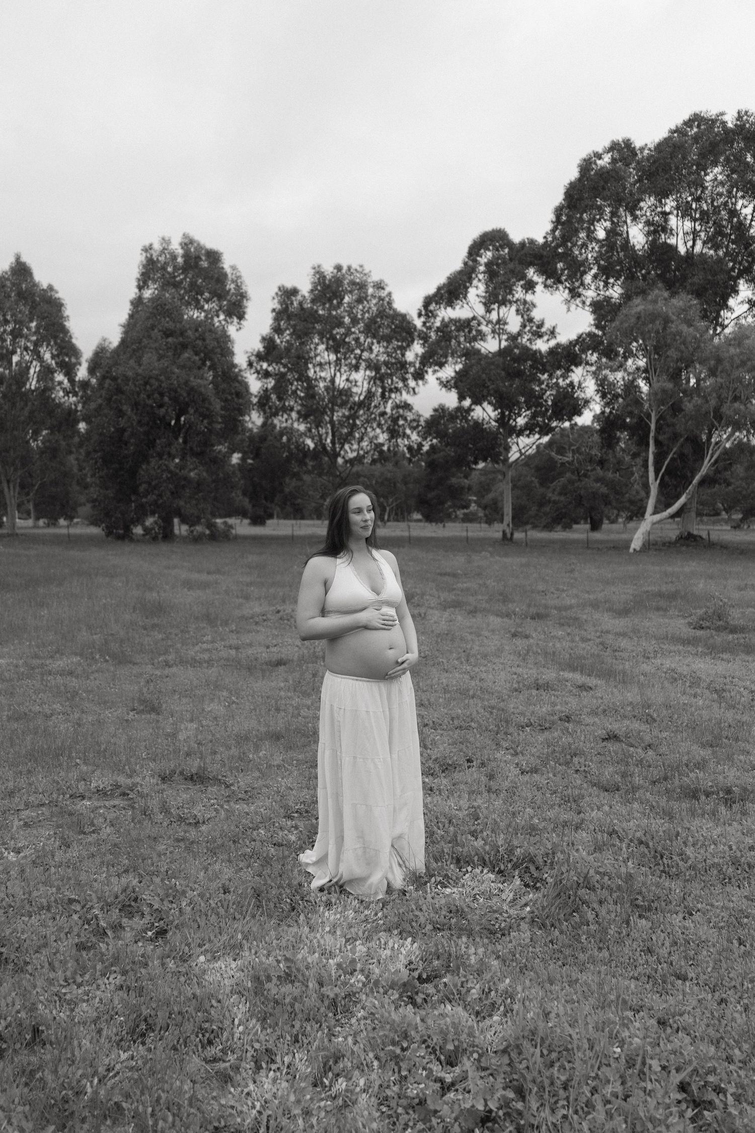 Black and white maternity photo of a pregnant woman in a white dress standing in an open park.