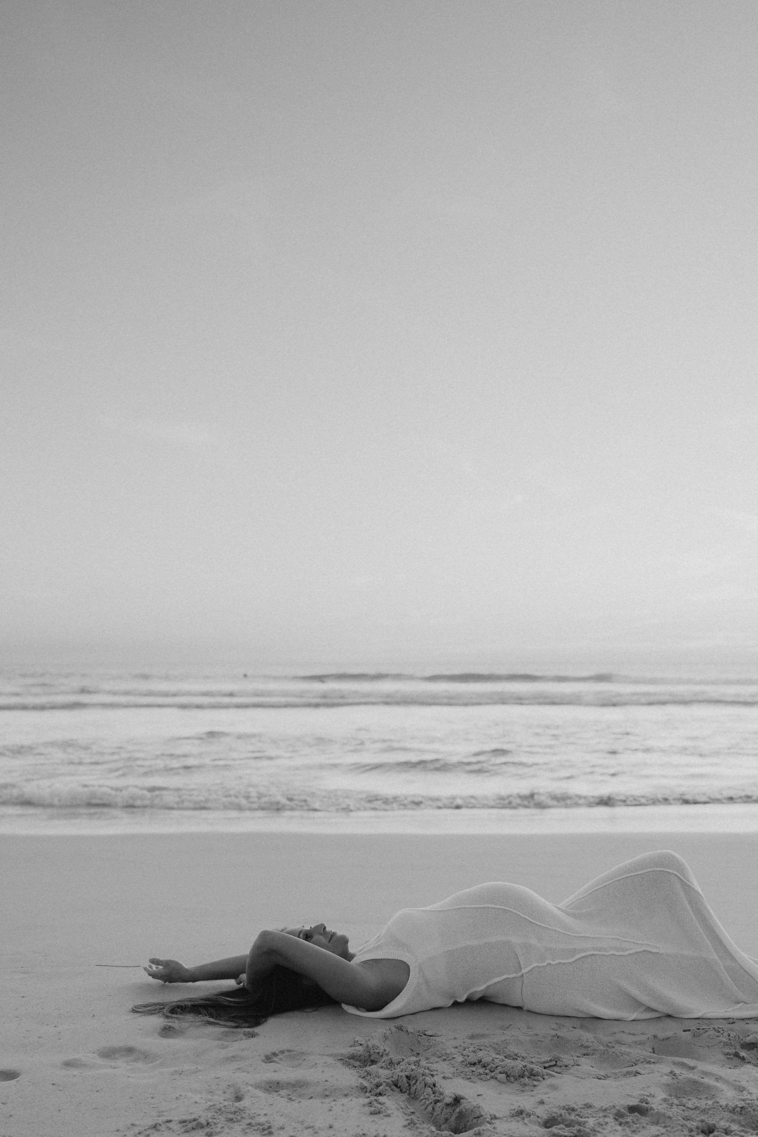 Pregnant woman in white dress lying on sandy beach with ocean waves in black and white photo.