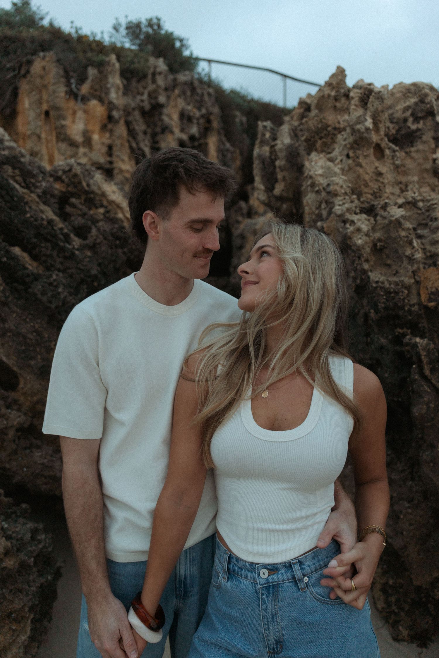 Smiling couple gazing at each other in front of rocky coastal cliffs, dressed in matching white and denim outfits.