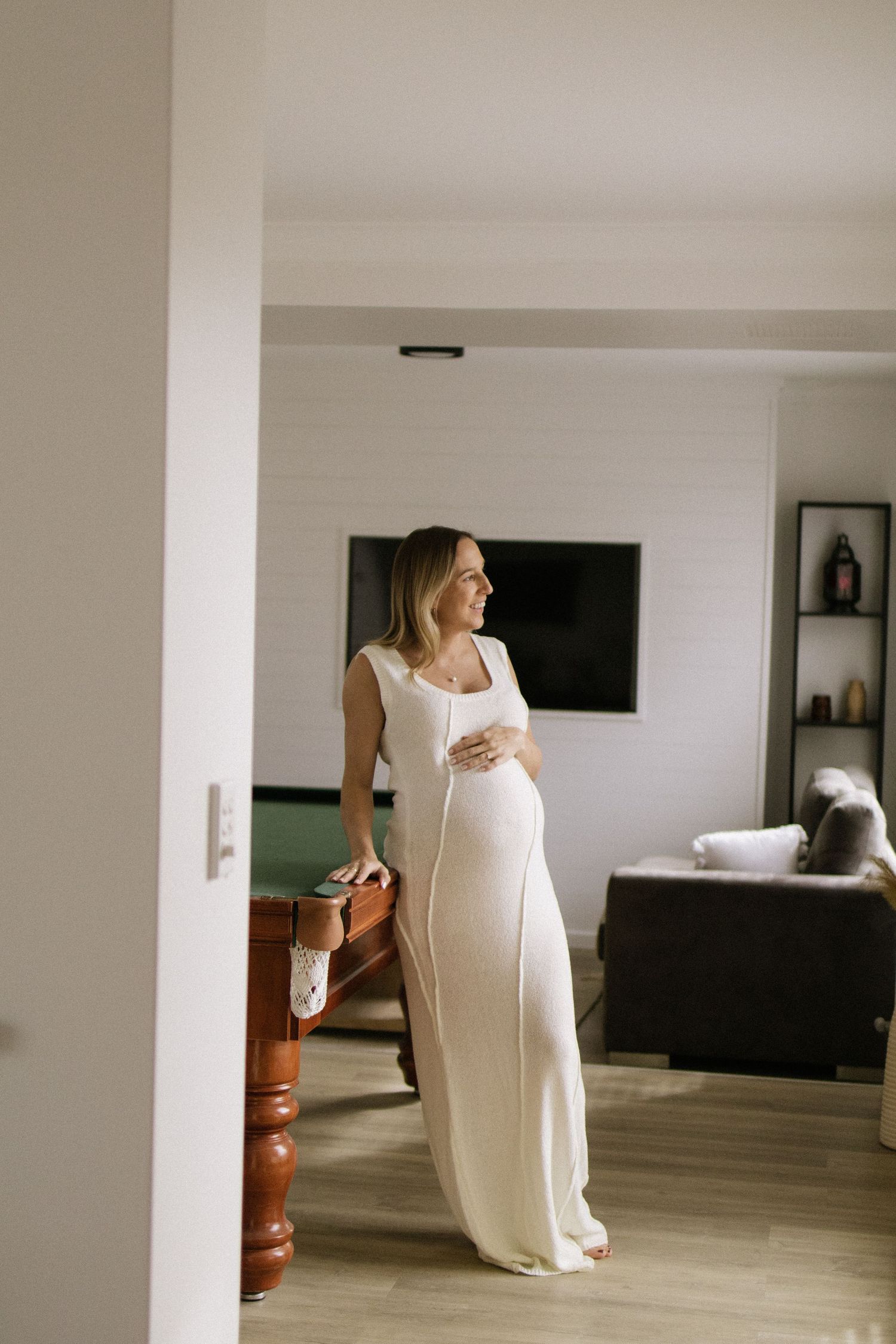 Smiling pregnant woman in white maxi dress standing in living room cradling her baby bump.