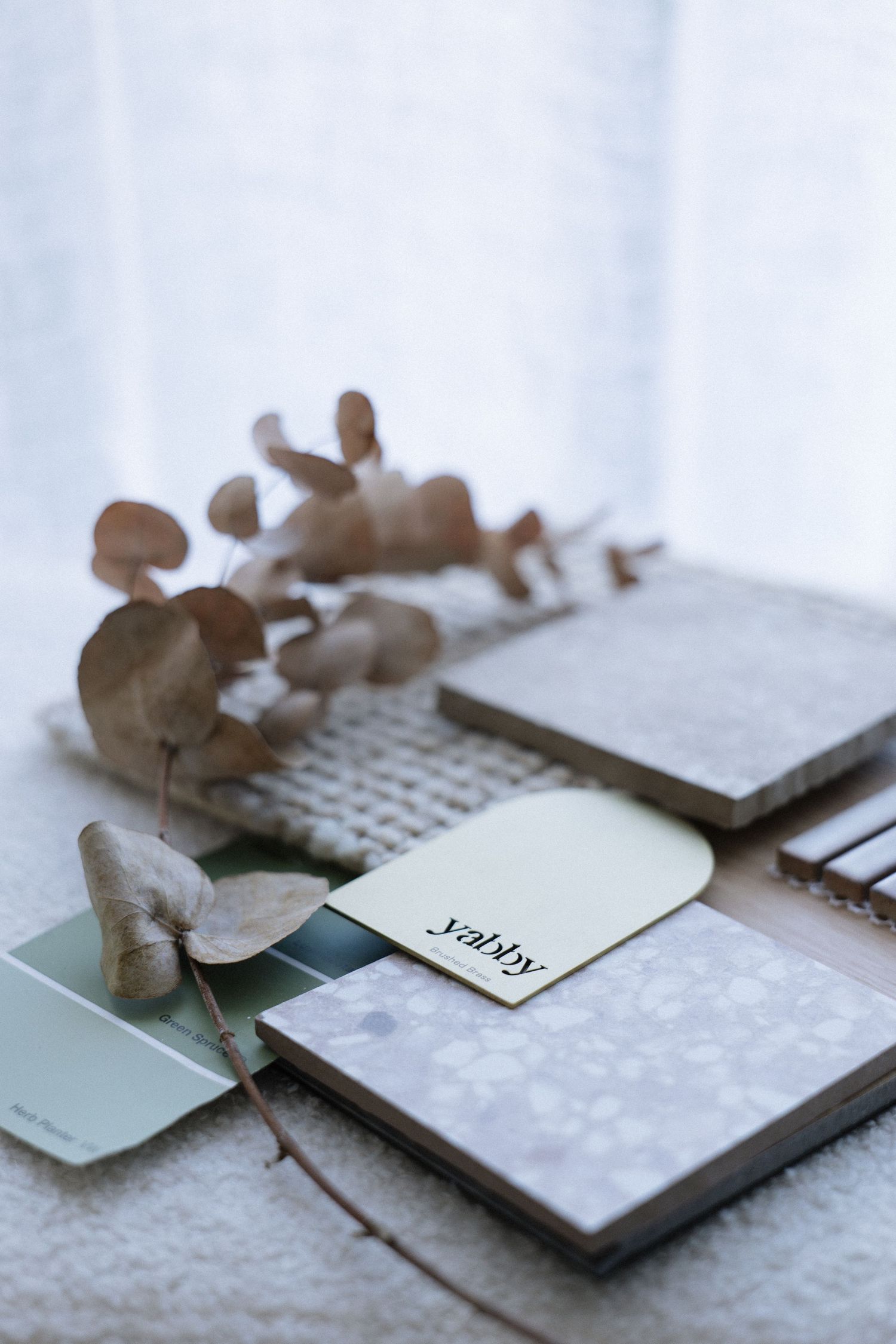 Interior design mood board with tile samples, paint swatches, and dried eucalyptus on white textured surface.