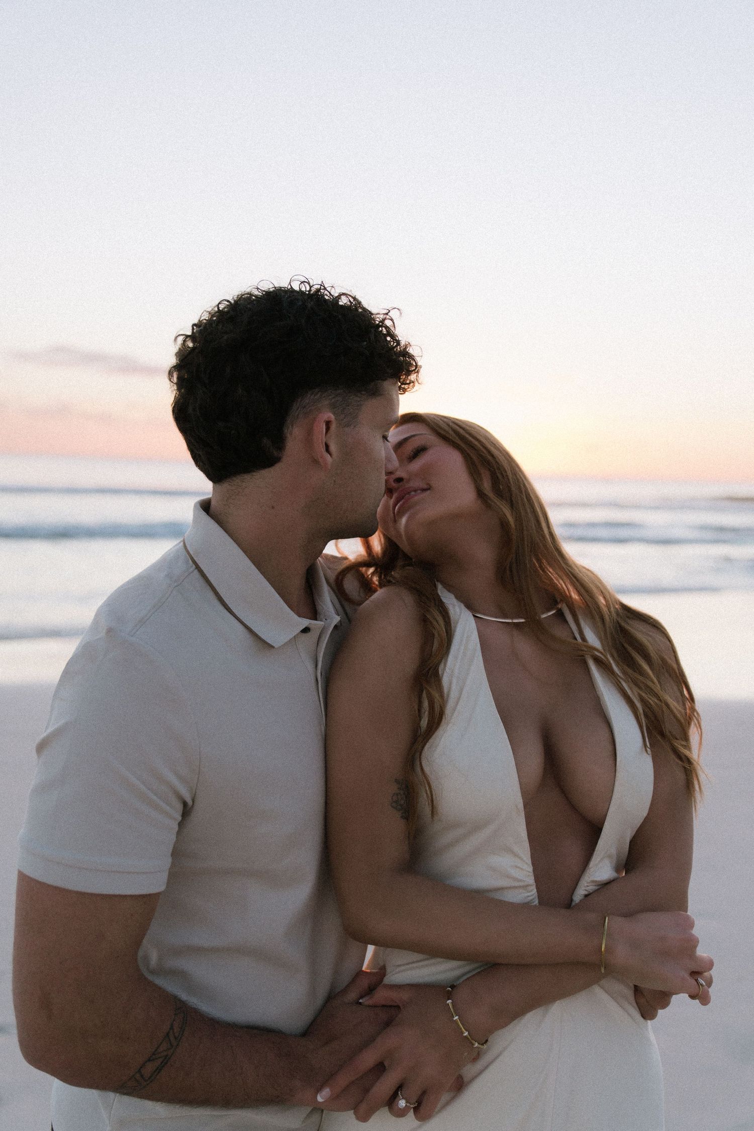 Couple sharing a tender kiss on beach at sunset, woman in white dress leaning into partner.