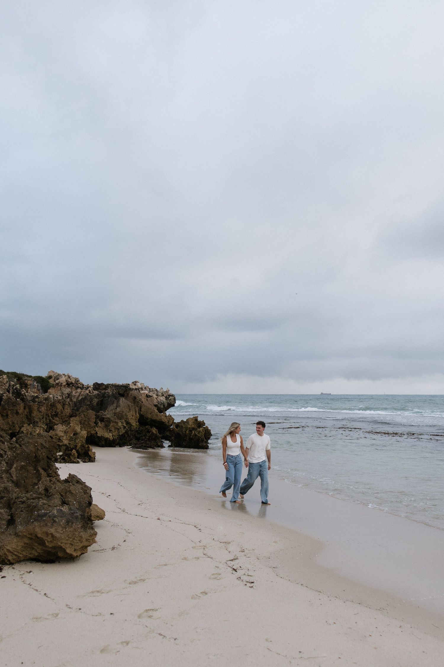 Couple walking hand in hand on sandy beach with rocky cliffs under cloudy sky.