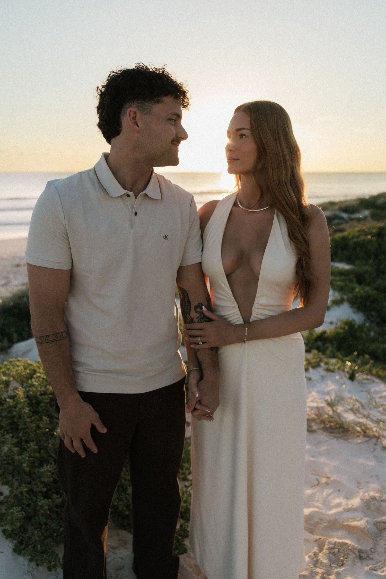 Couple standing together at sunset on beach dunes, woman in elegant white halter dress.