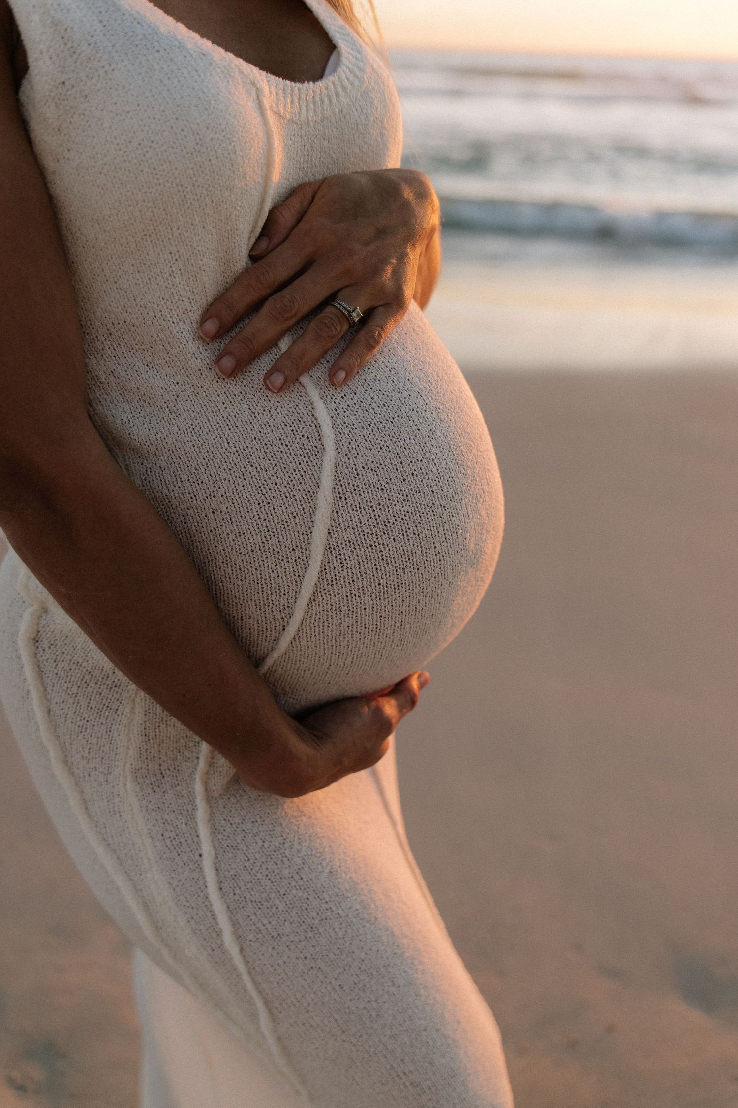 Close-up of pregnant woman in white knit dress holding baby bump on beach at sunset.