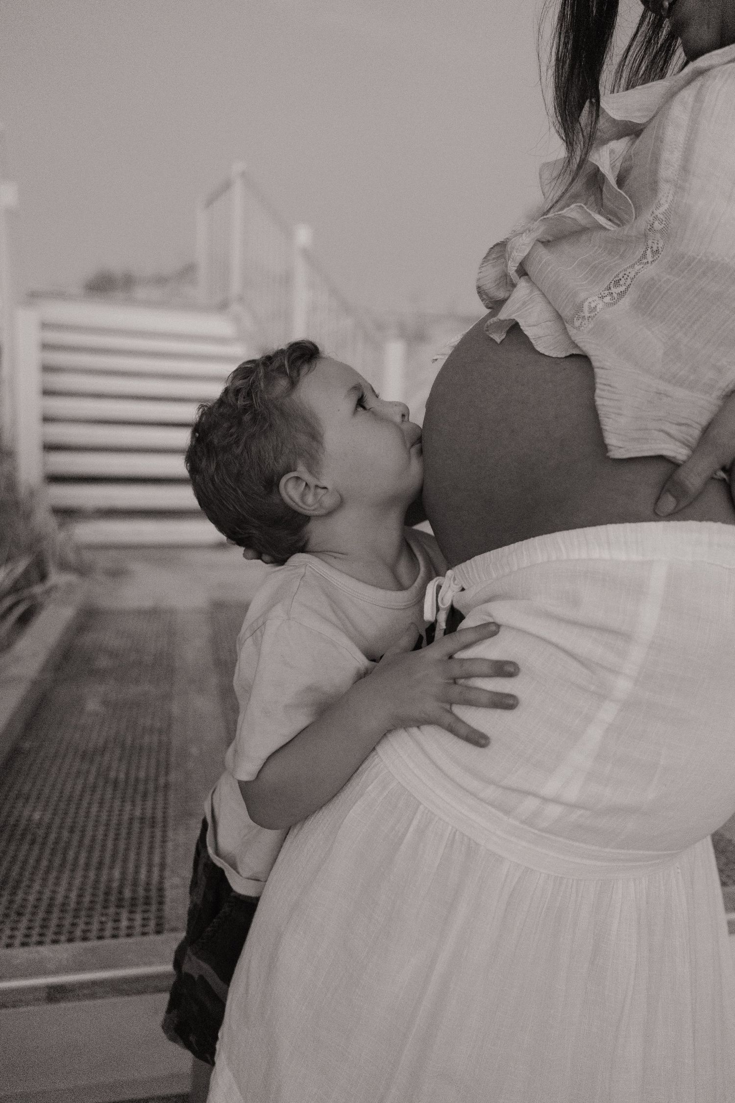 Young child kissing pregnant mother's baby bump in black and white photo.