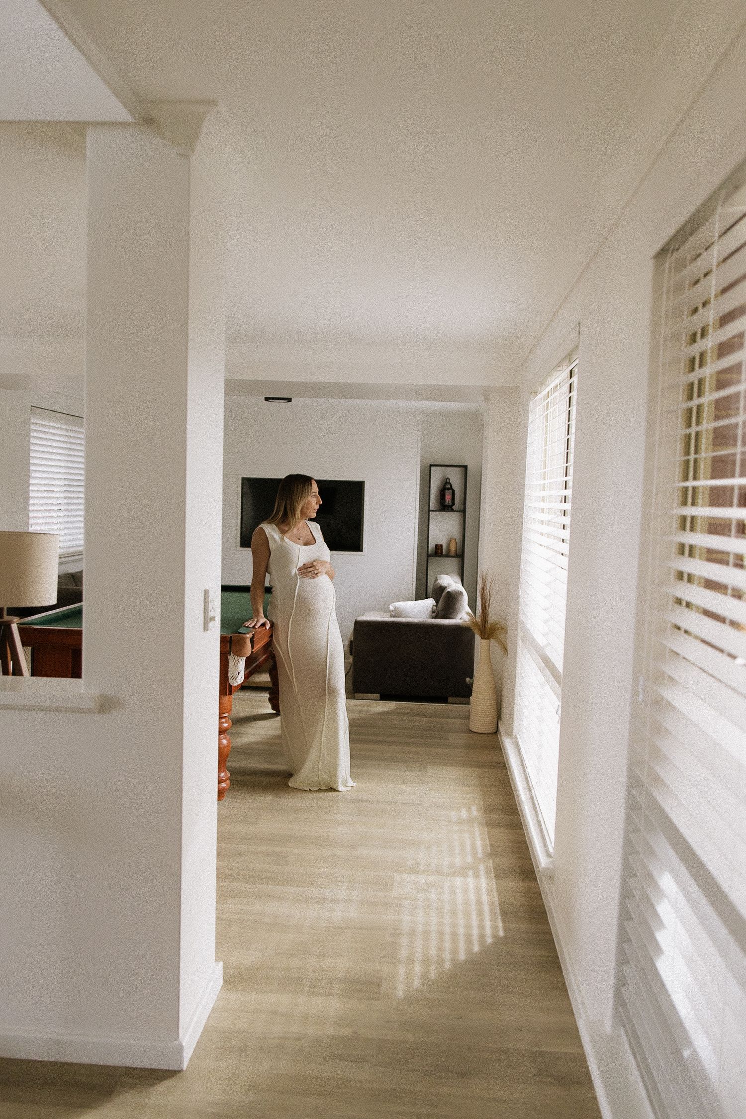 Pregnant woman in white maxi dress stands in sunlit hallway near window blinds.