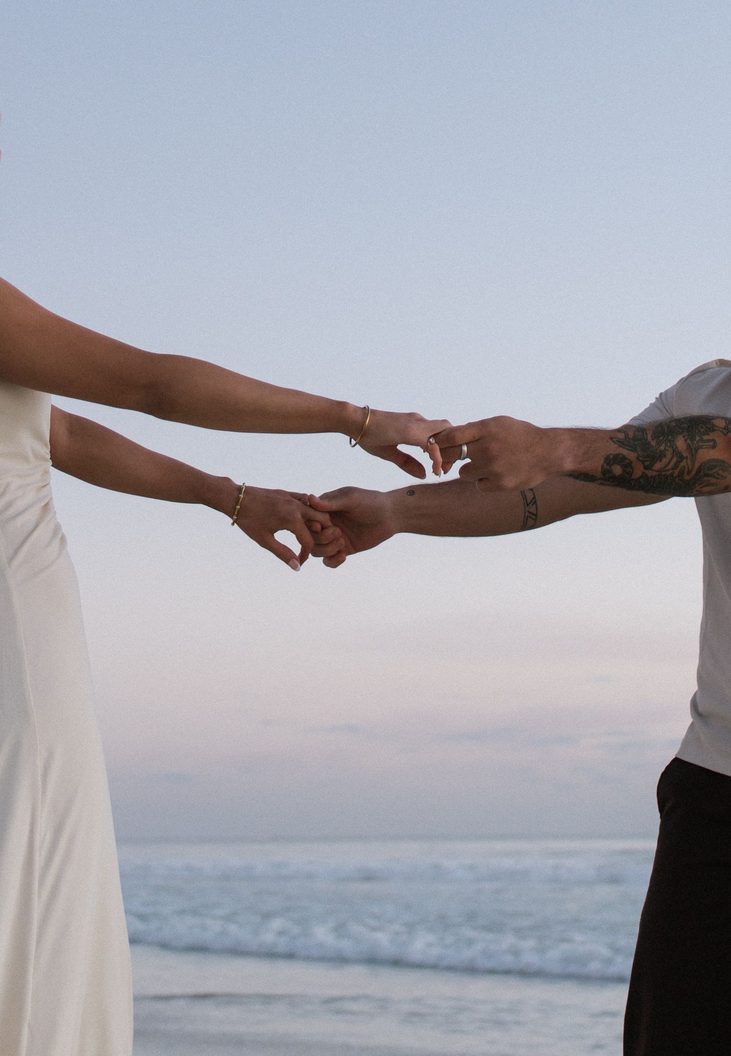 Couple holding hands at beach sunset, bride in white dress with tattooed groom.