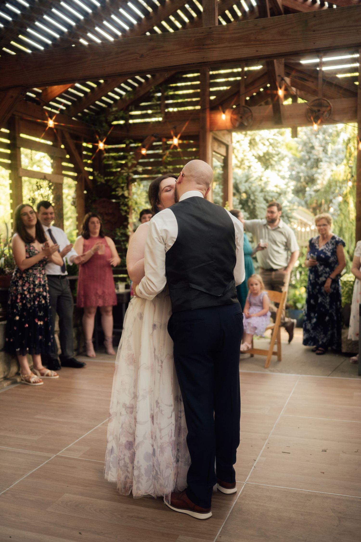 Bride and groom share first dance under rustic wooden pavilion with string lights at outdoor wedding reception.