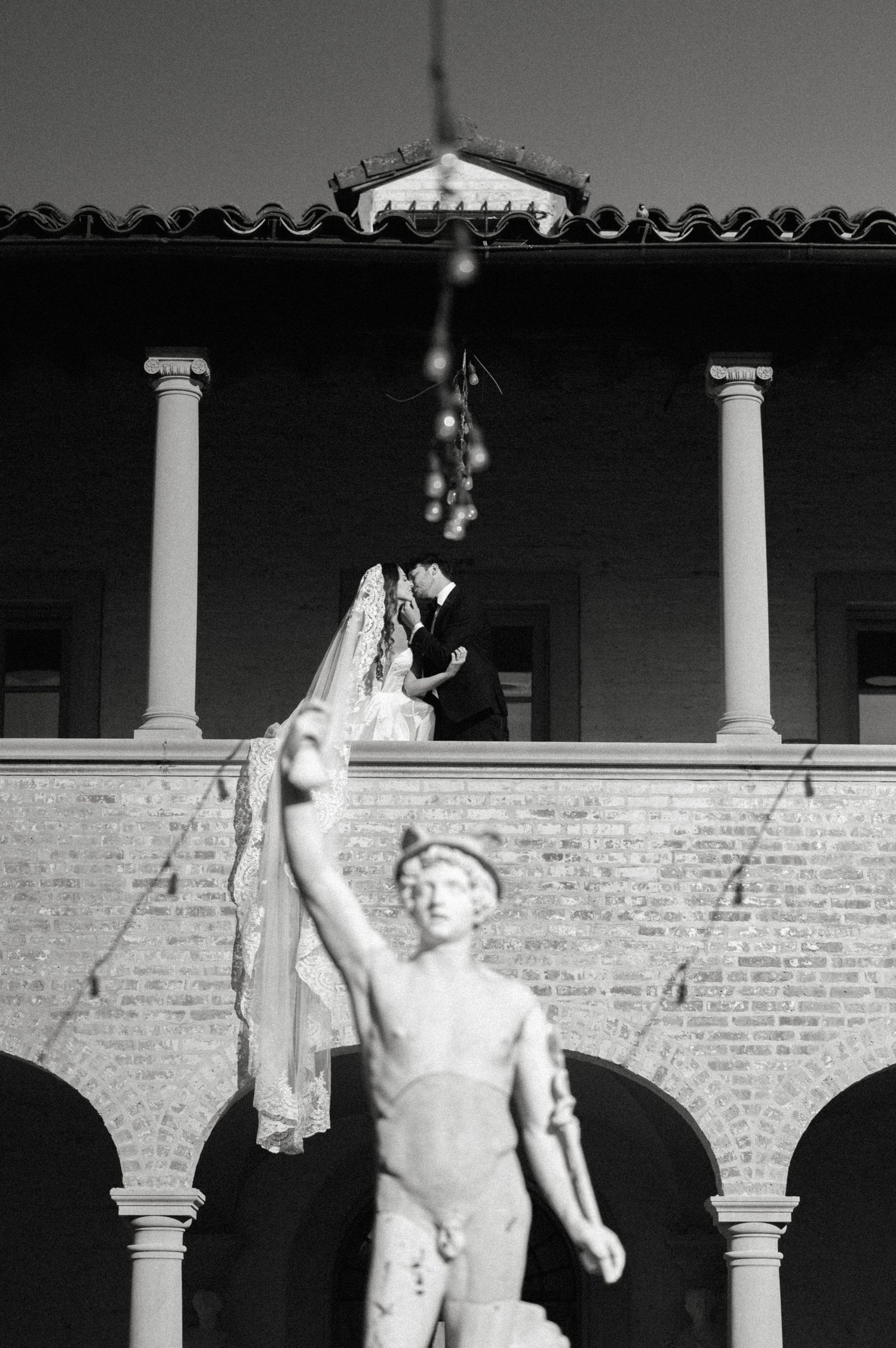 A wedding couple kisses on a balcony above a classical statue at an elegant brick venue in black and white.