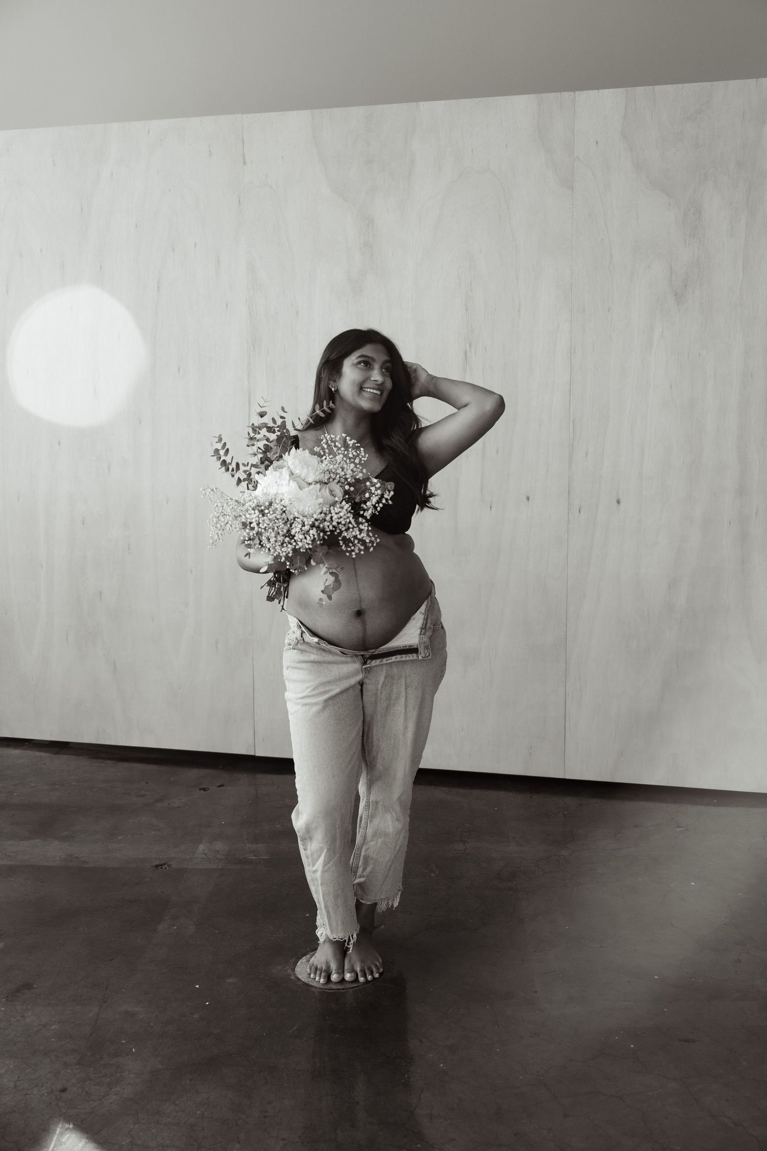 Black and white photo of smiling pregnant woman holding flowers in white pants.