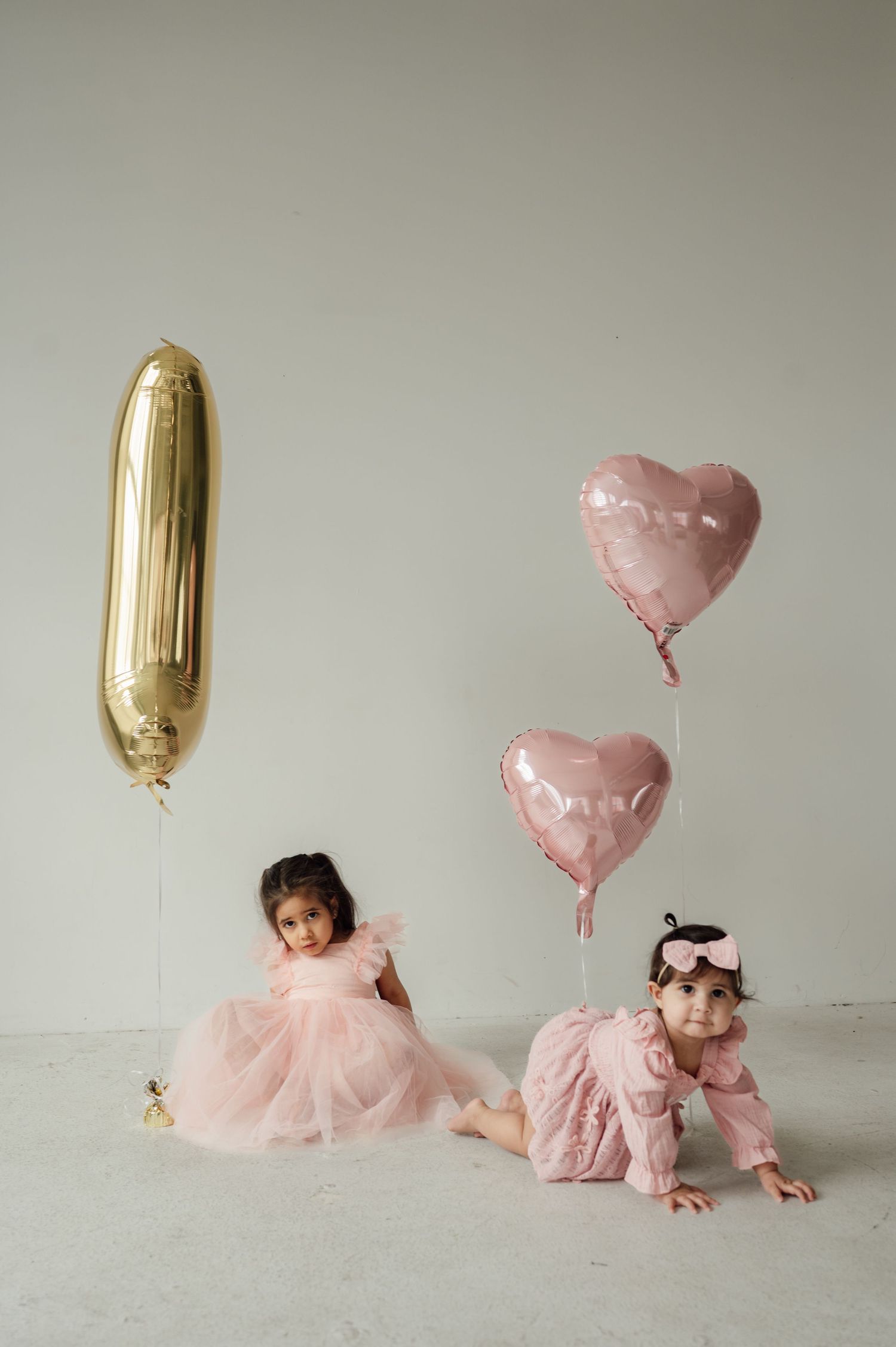 Two young girls in pink dresses posing with heart and number one balloons for first birthday.