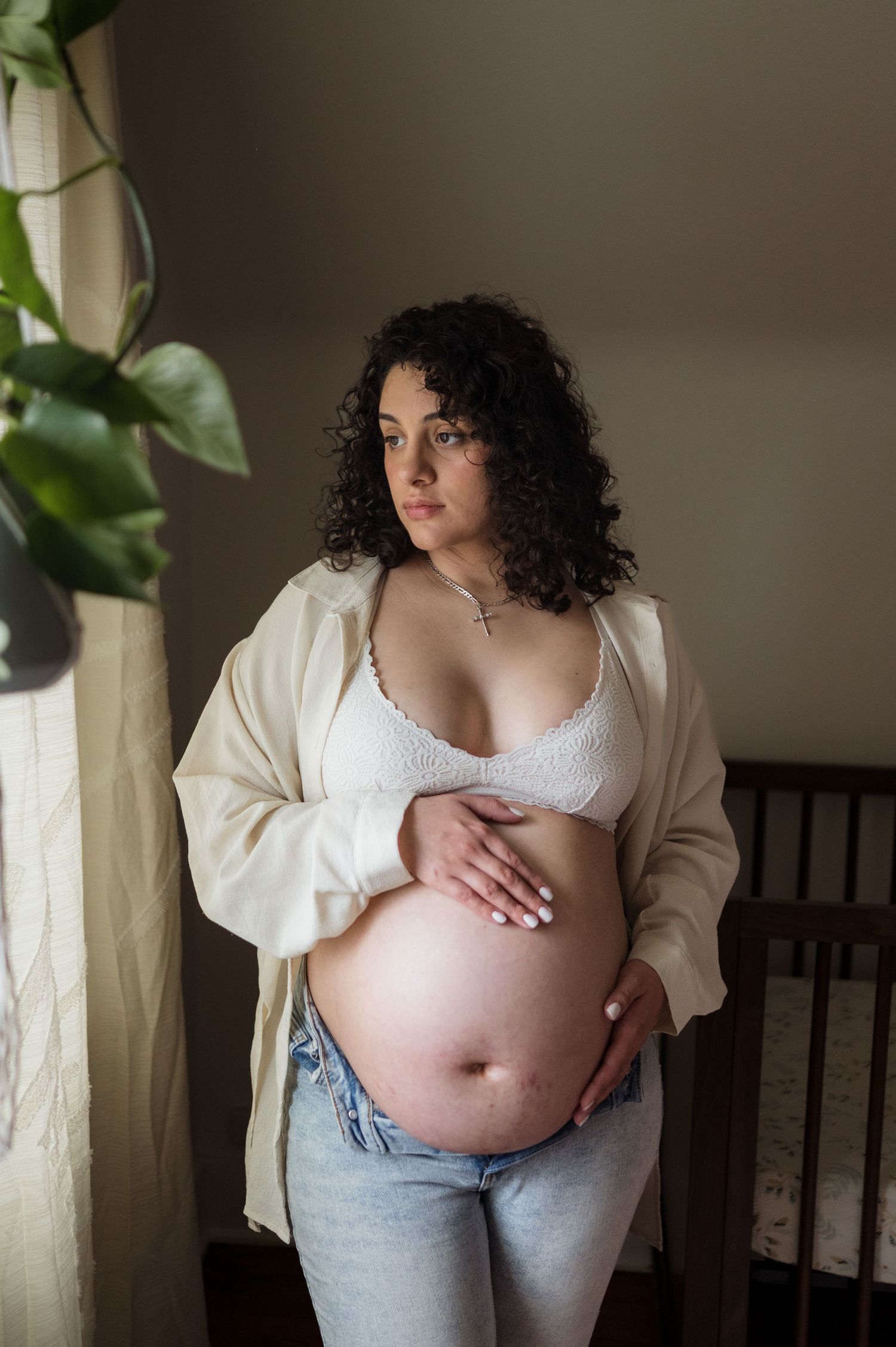 Pregnant woman in white lace bralette and jeans cradling her baby bump while standing near a window in soft natural light.