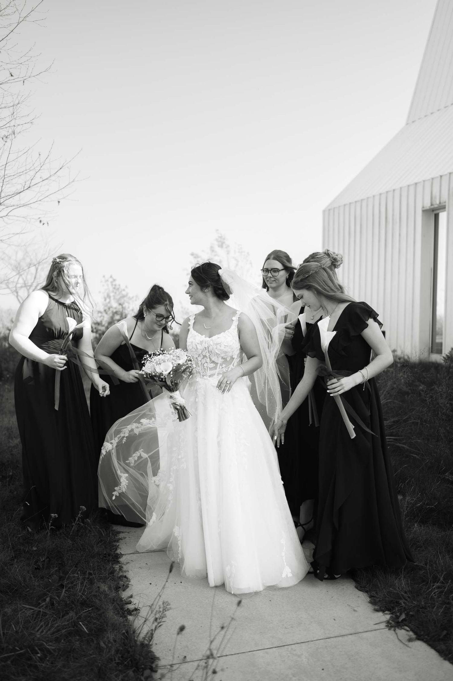 Black and white photo of bride in lace gown surrounded by bridesmaids in dark dresses outdoors.