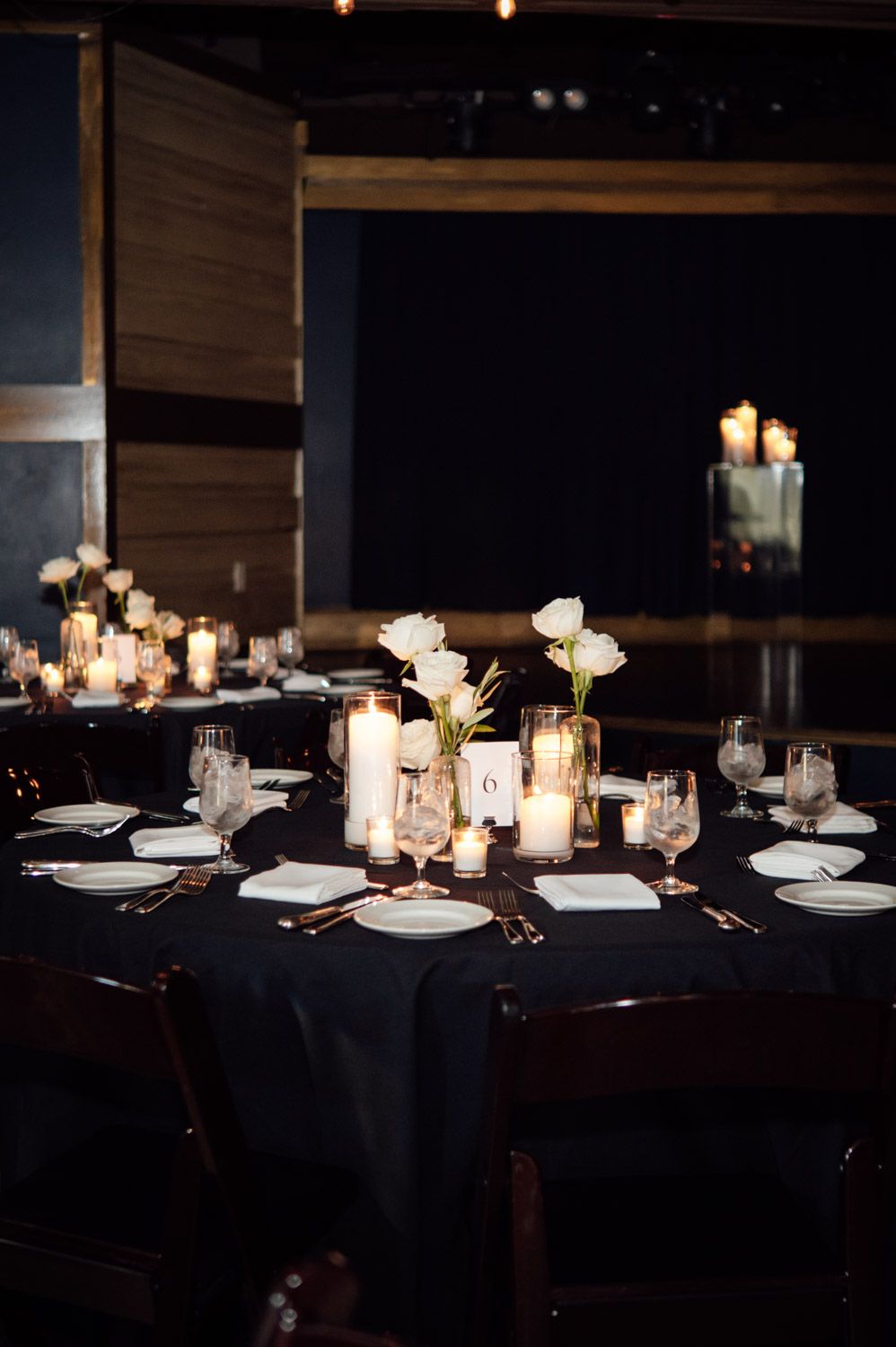 Elegant dark wedding reception table with white roses, pillar candles, and glassware centerpieces.