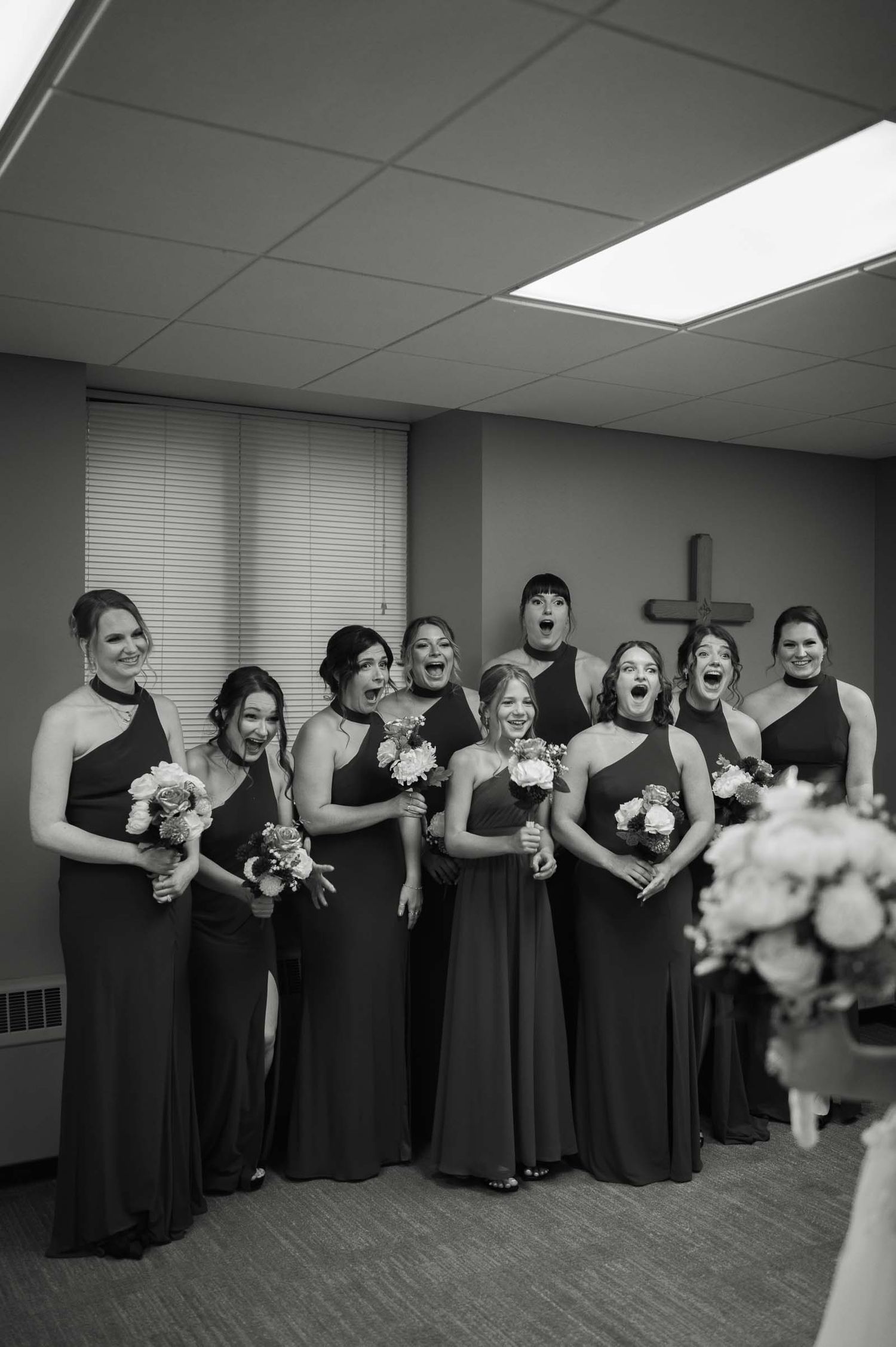 Bridesmaids in black gowns react with excitement and joy while holding bouquets inside a church room.
